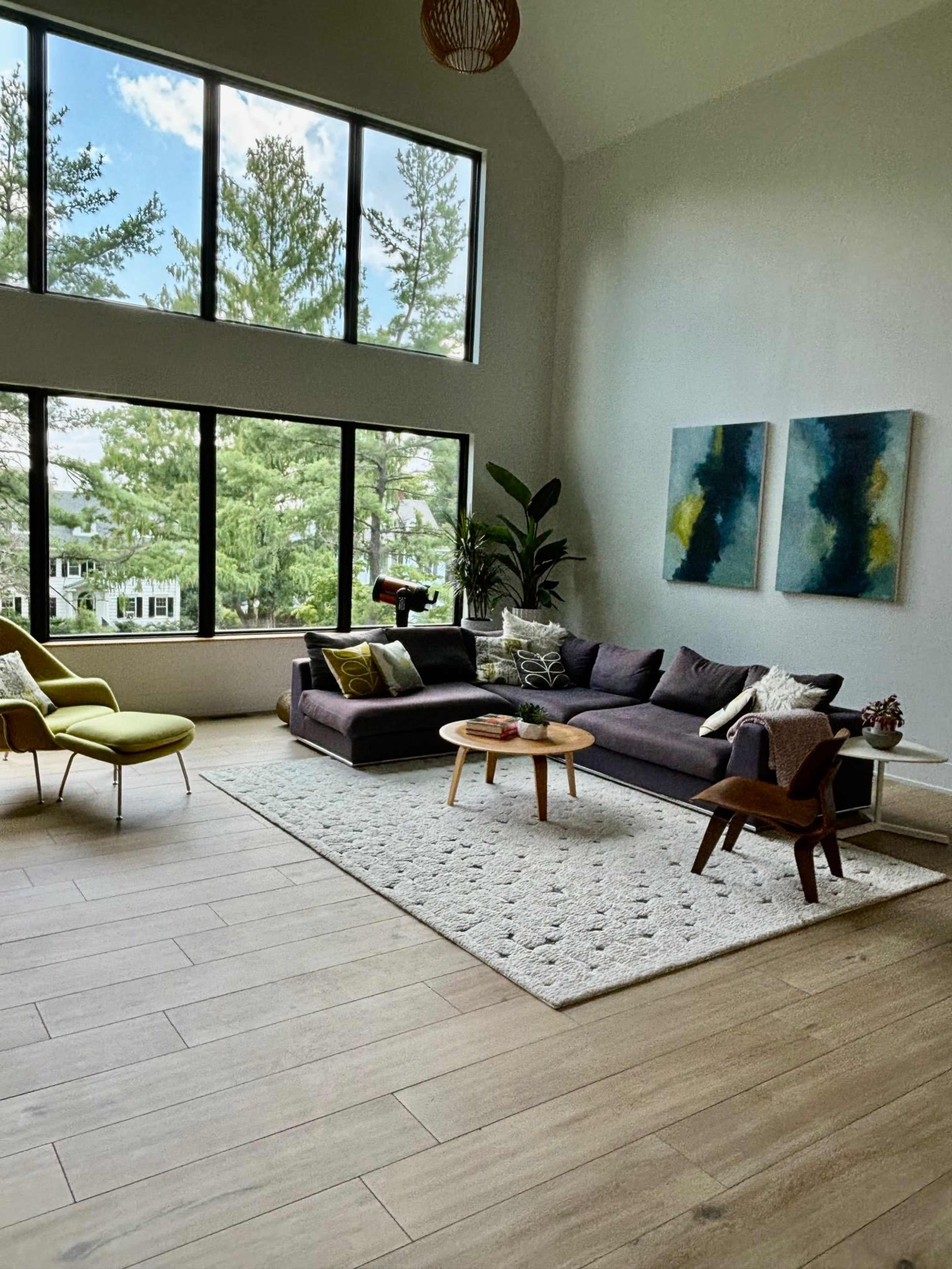 A spacious living room features large windows, a dark sofa, a round coffee table, and modern art on the walls.