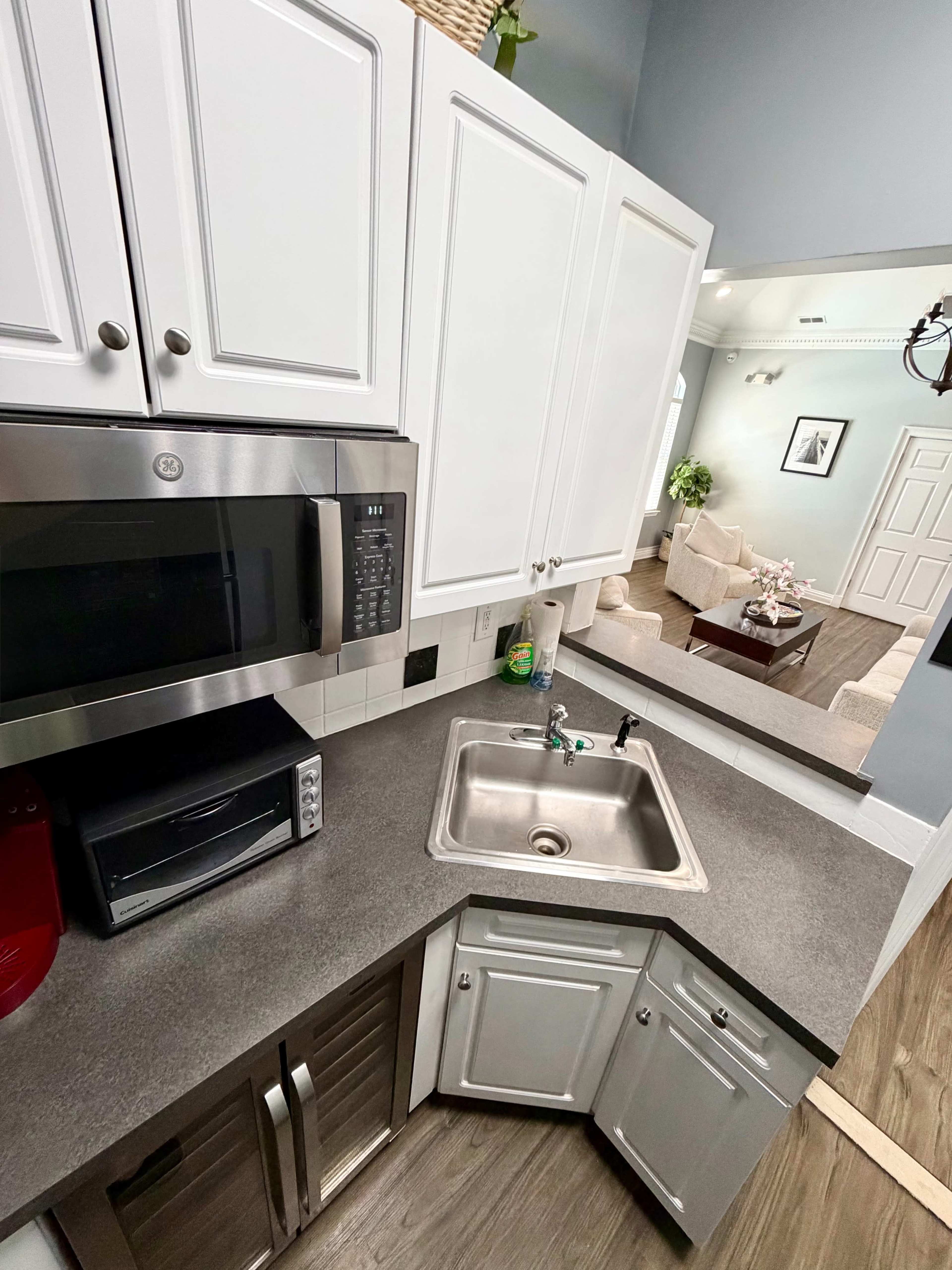 The image shows a corner kitchen with a stainless steel sink, a microwave, and gray countertops, while a living area is visible through an open doorway.