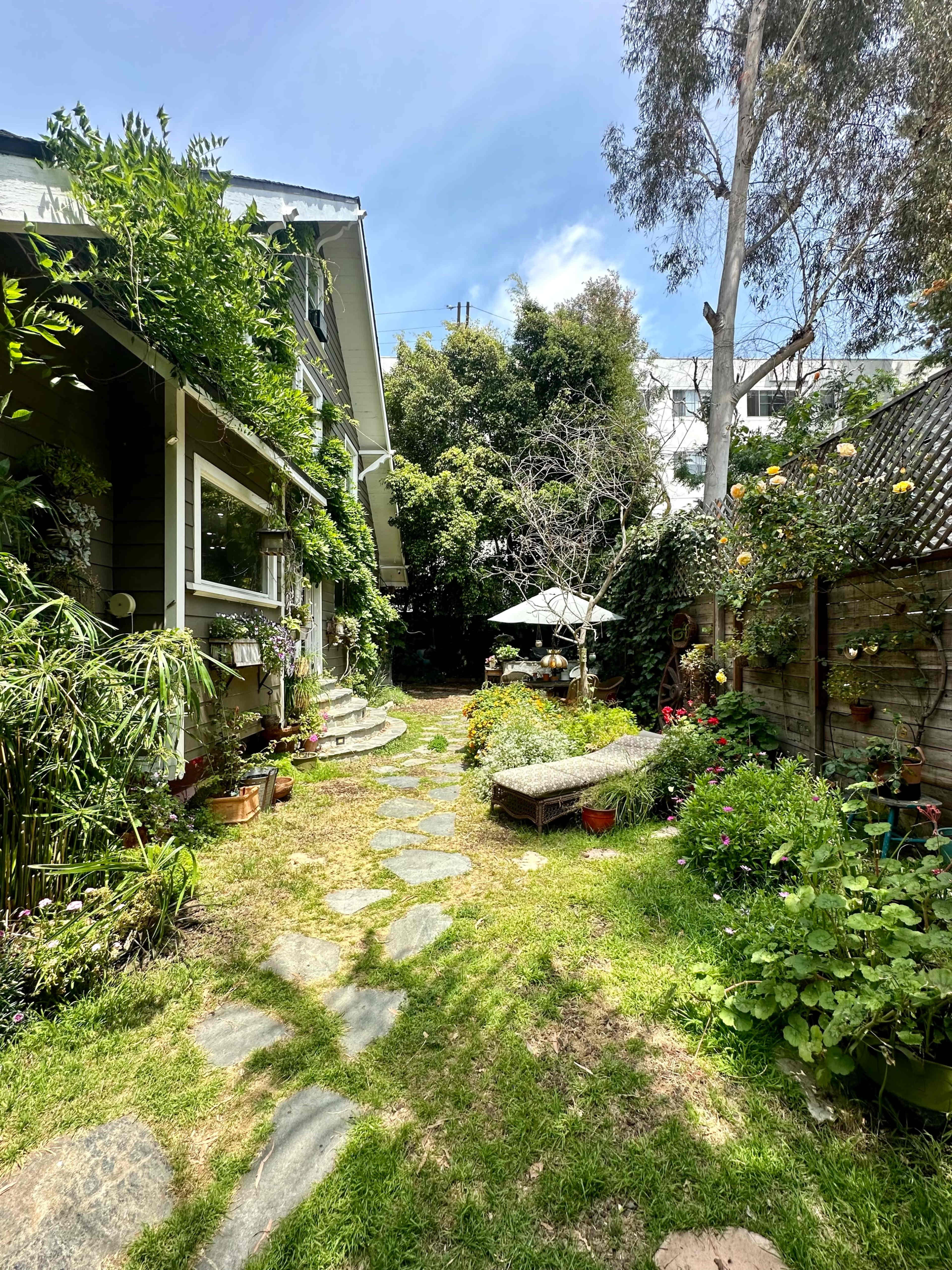 A stone pathway leads through a garden filled with various plants and flowers, alongside a house and an umbrella-covered seating area.