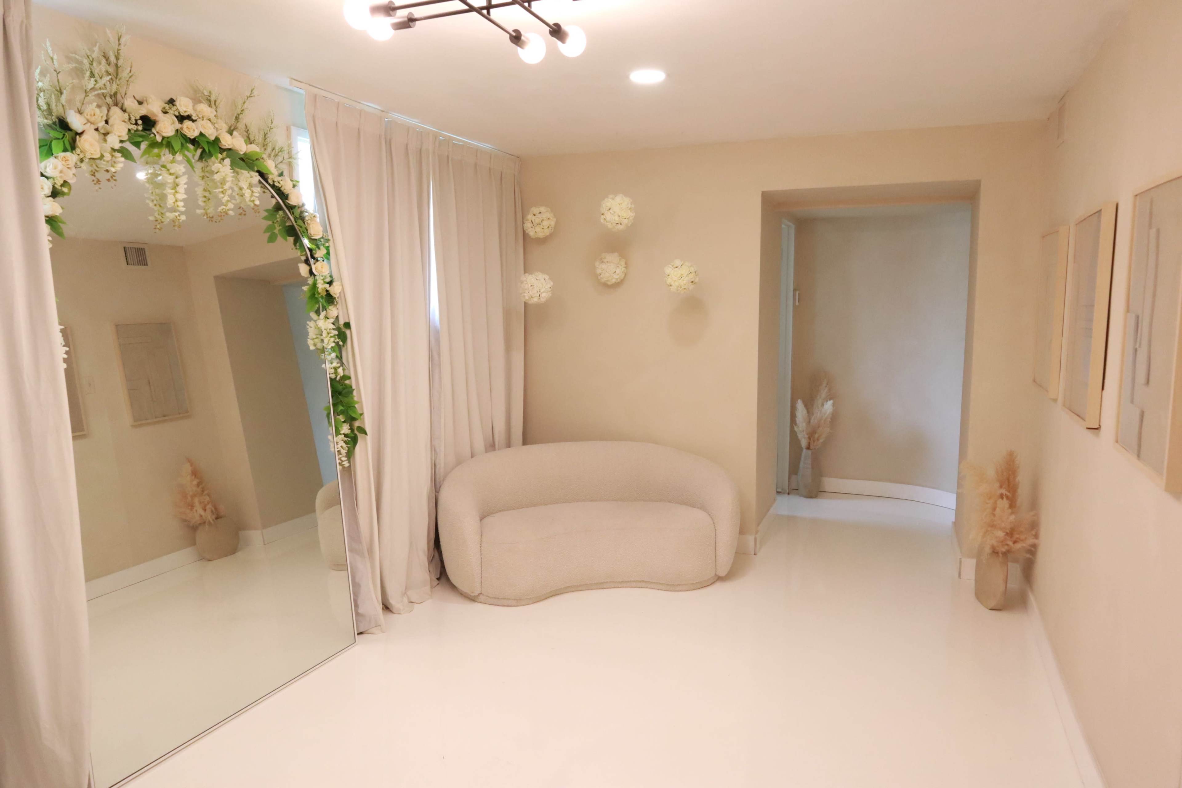 The image shows a minimalistic interior space featuring a curved beige couch, a large mirror with floral decor, and several wall-mounted decorations, all set against a light-colored wall.