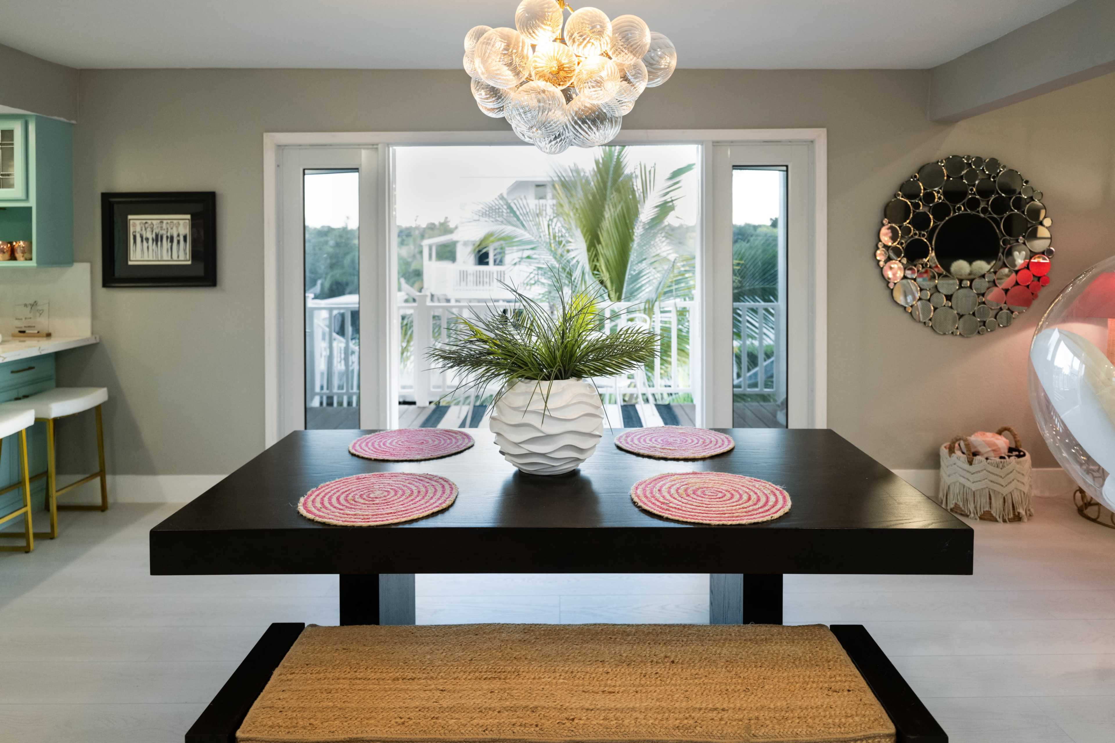 A modern dining area features a black table with a white vase of greenery, set with round placemats, and a striking chandelier above it.