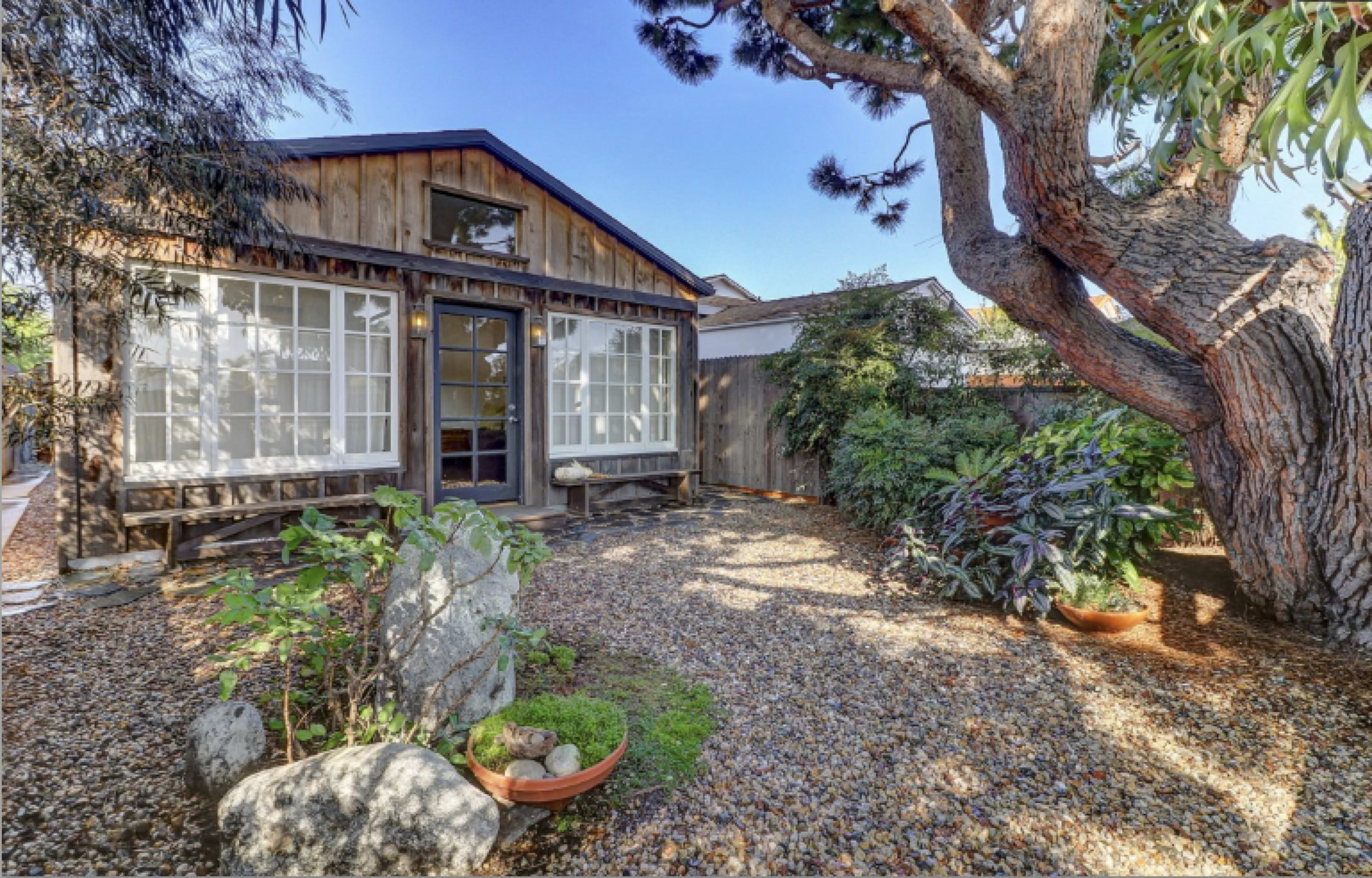 A wooden cabin with large windows is surrounded by a landscaped garden featuring gravel pathways and various plants.