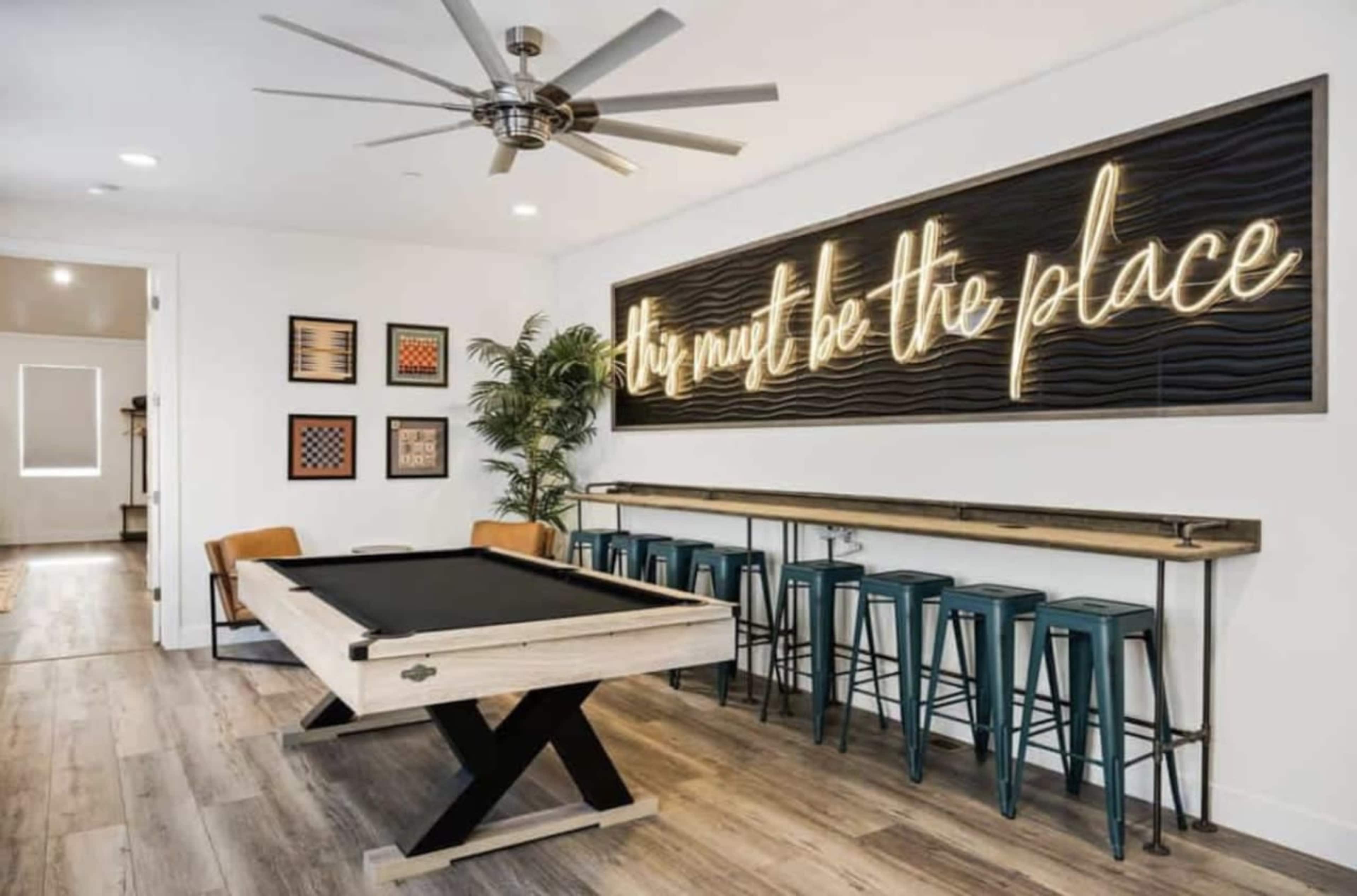 The image features a game room with a pool table, a neon sign on the wall, and seating at a bar-style table.
