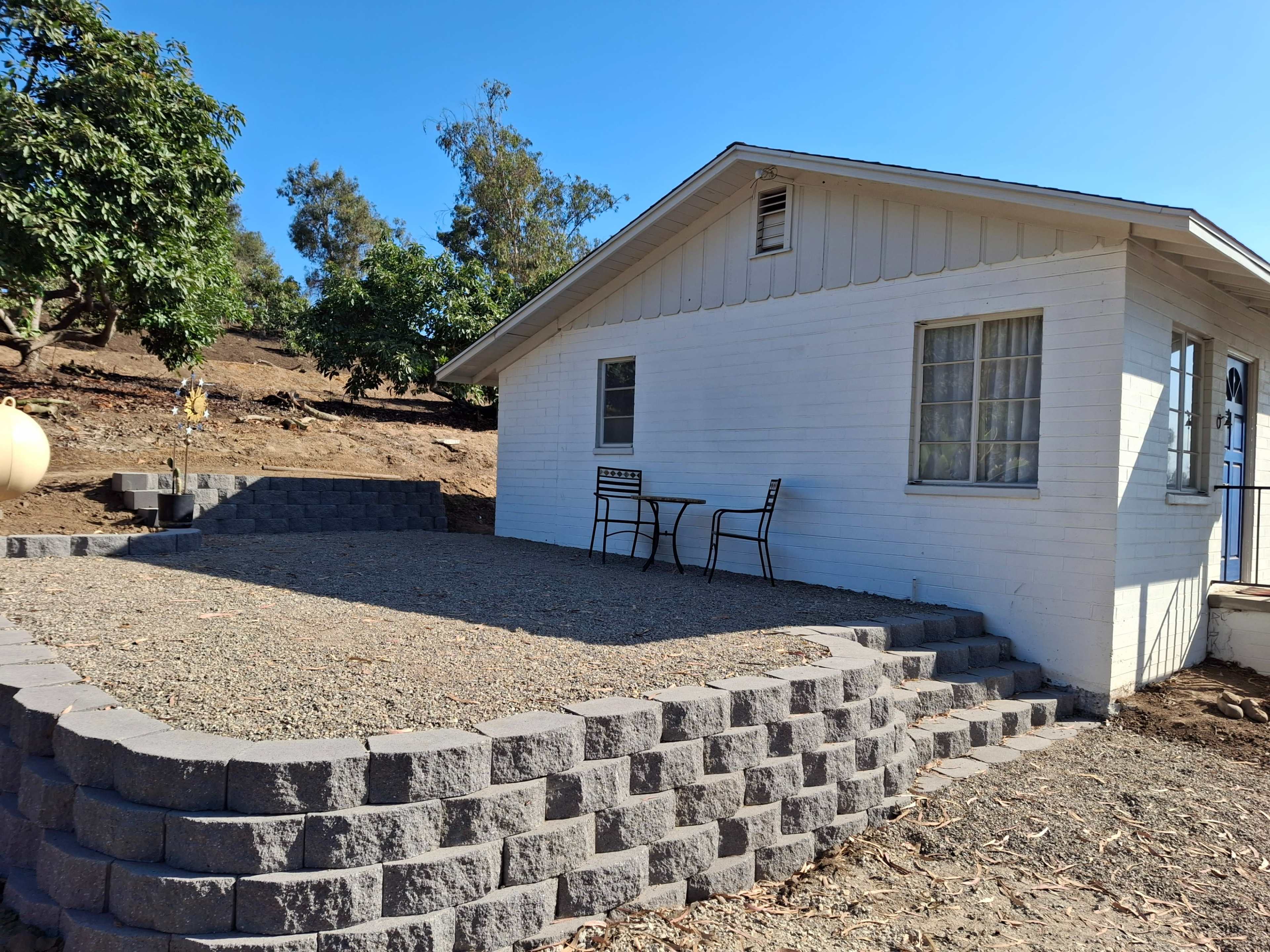 A small white house with a front porch area features a table and chairs, situated on landscaped gravel with a stone retaining wall.