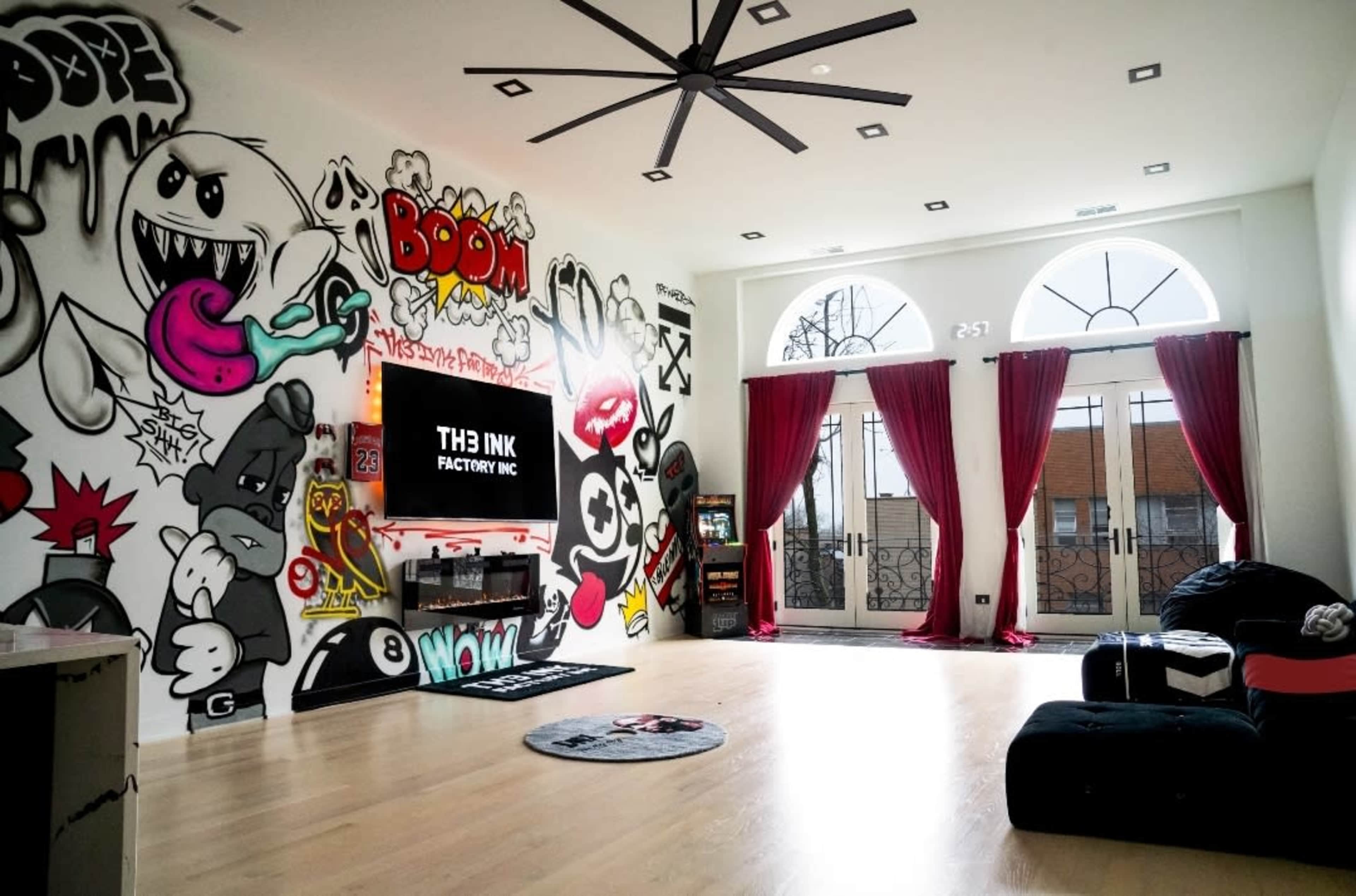 The room features a wall covered in colorful graffiti, a large television mounted on a sleek unit, and two red curtains framing a set of arched windows.