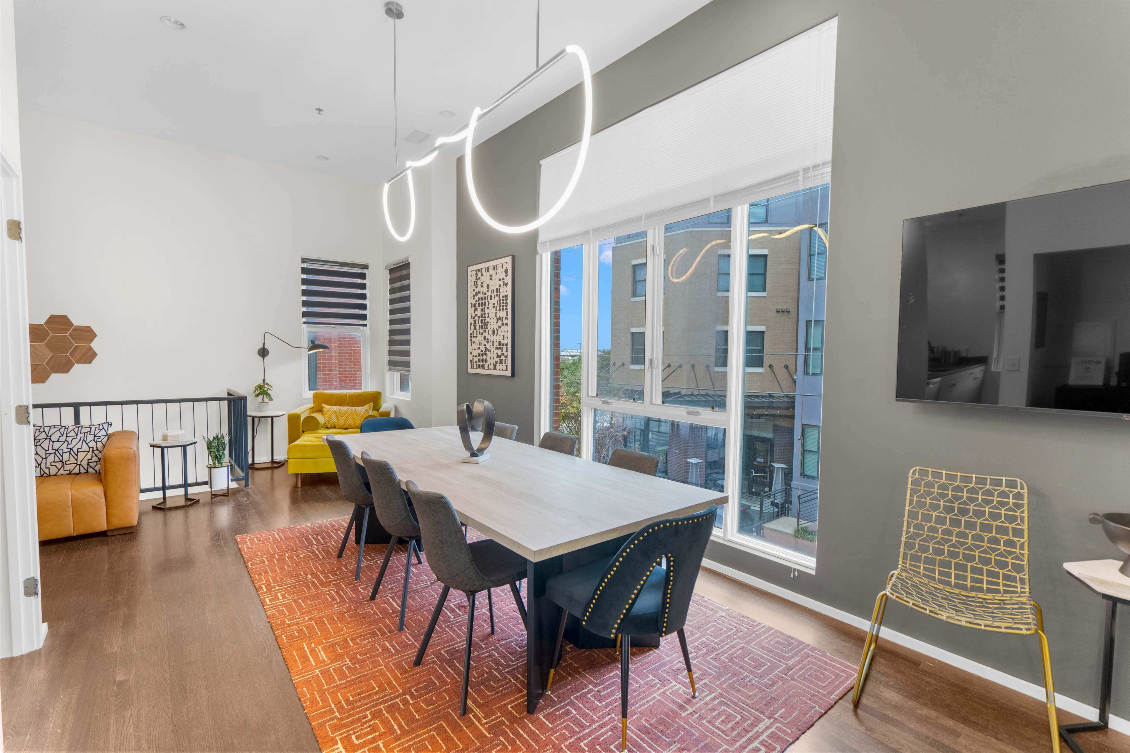 A modern dining area features a sleek table with black chairs, illuminated by contemporary light fixtures, adjacent to a living space with a yellow sofa and large windows.