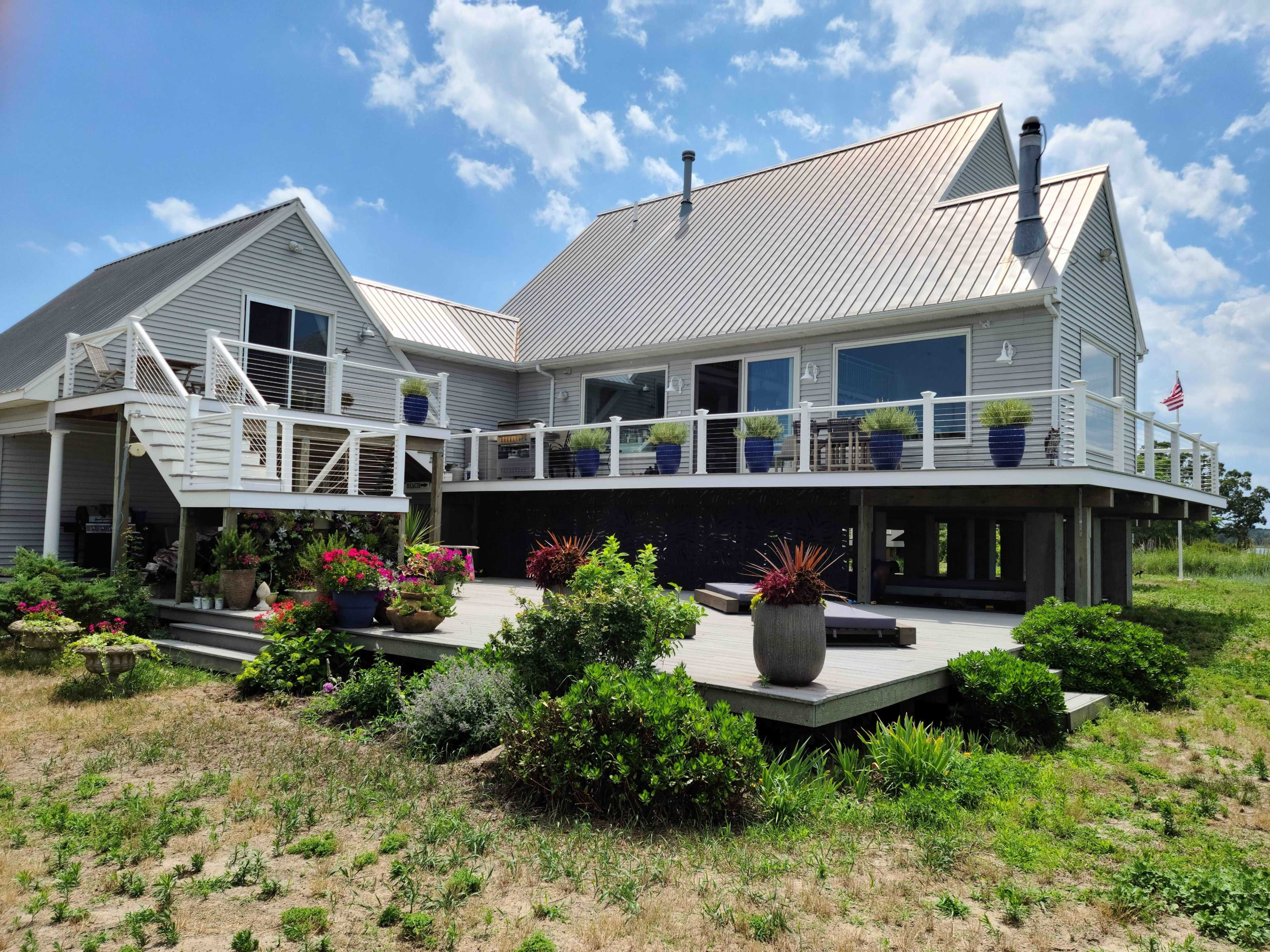 A large, modern house with a metal roof and a wraparound deck is surrounded by green grass and potted plants.