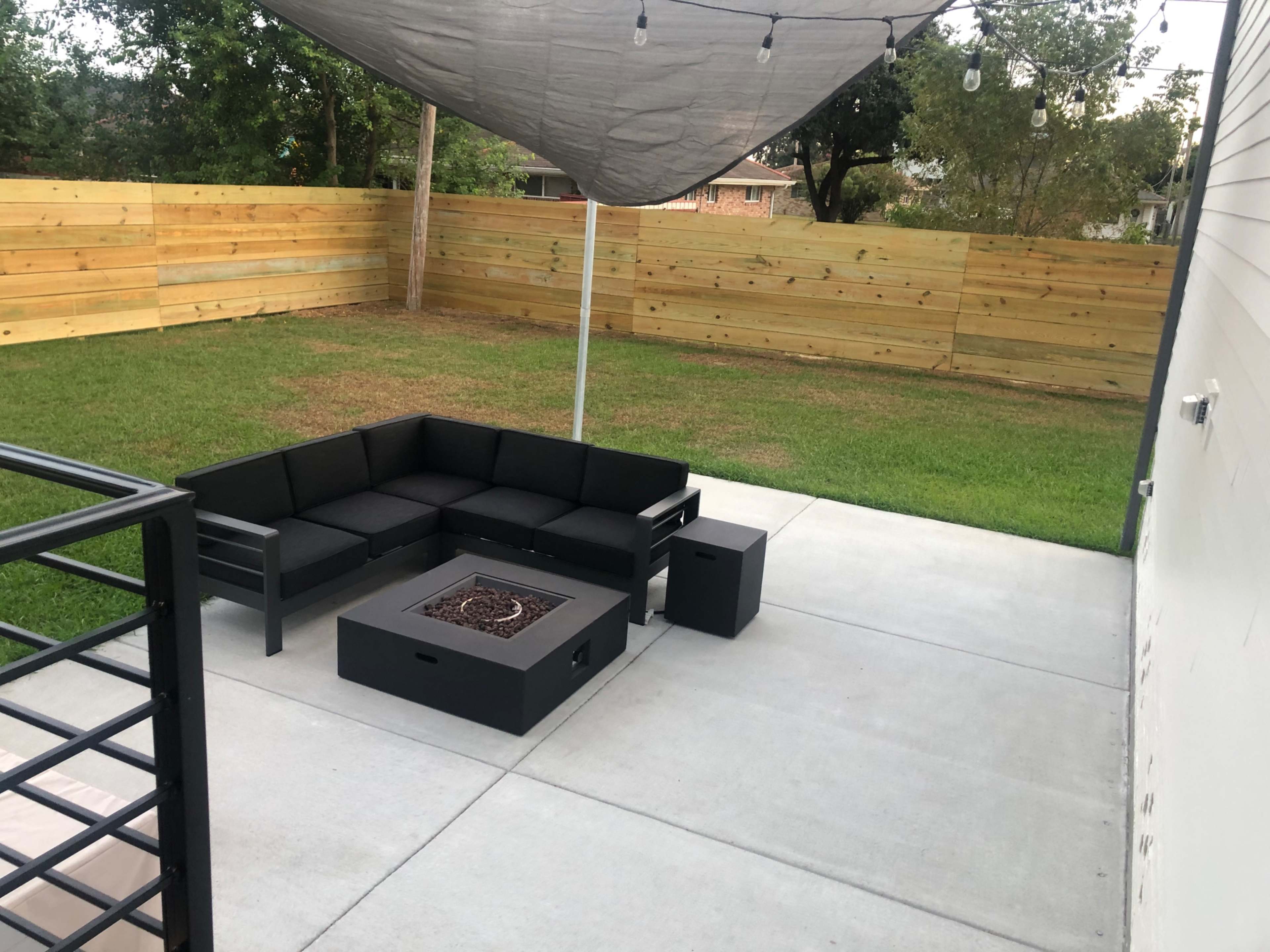 A modern outdoor seating area features a black sectional sofa and a fire pit under a shaded canopy on a concrete patio, surrounded by a wooden fence and grass.