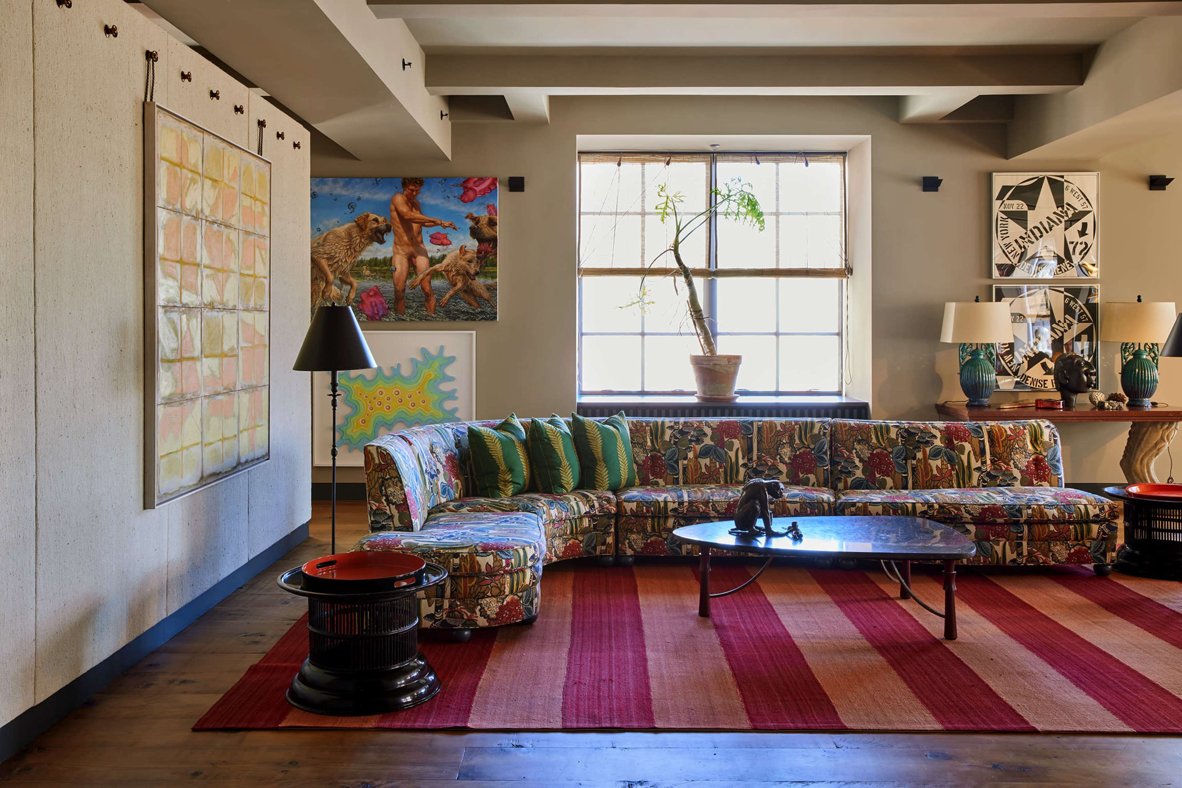 The room features a colorful patterned sectional sofa, a round coffee table, and various pieces of artwork on the walls, with a large window providing natural light.