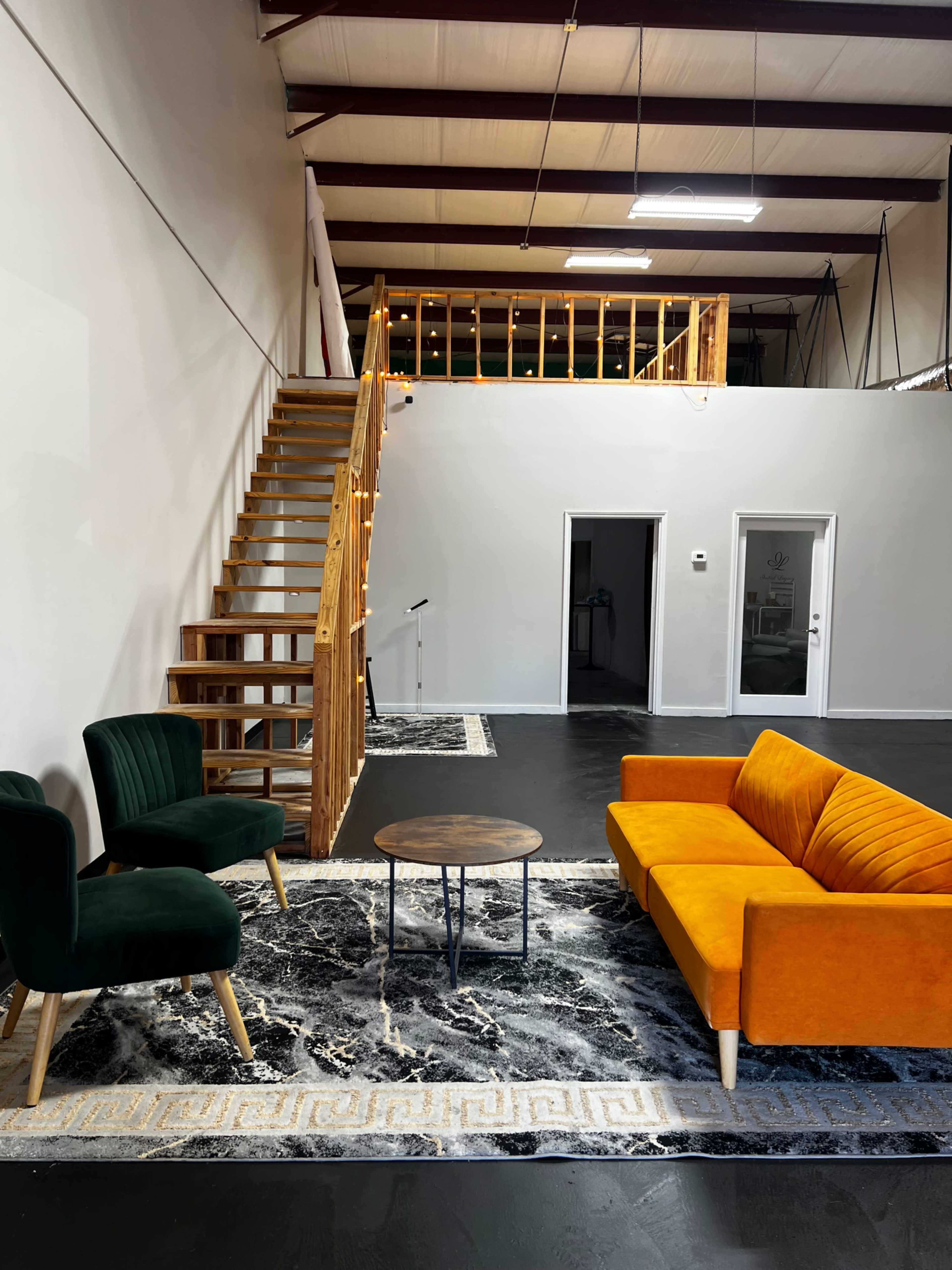 The image shows a modern interior with a vibrant orange sofa, green armchairs, a round coffee table, and a wooden staircase leading to an upper level.