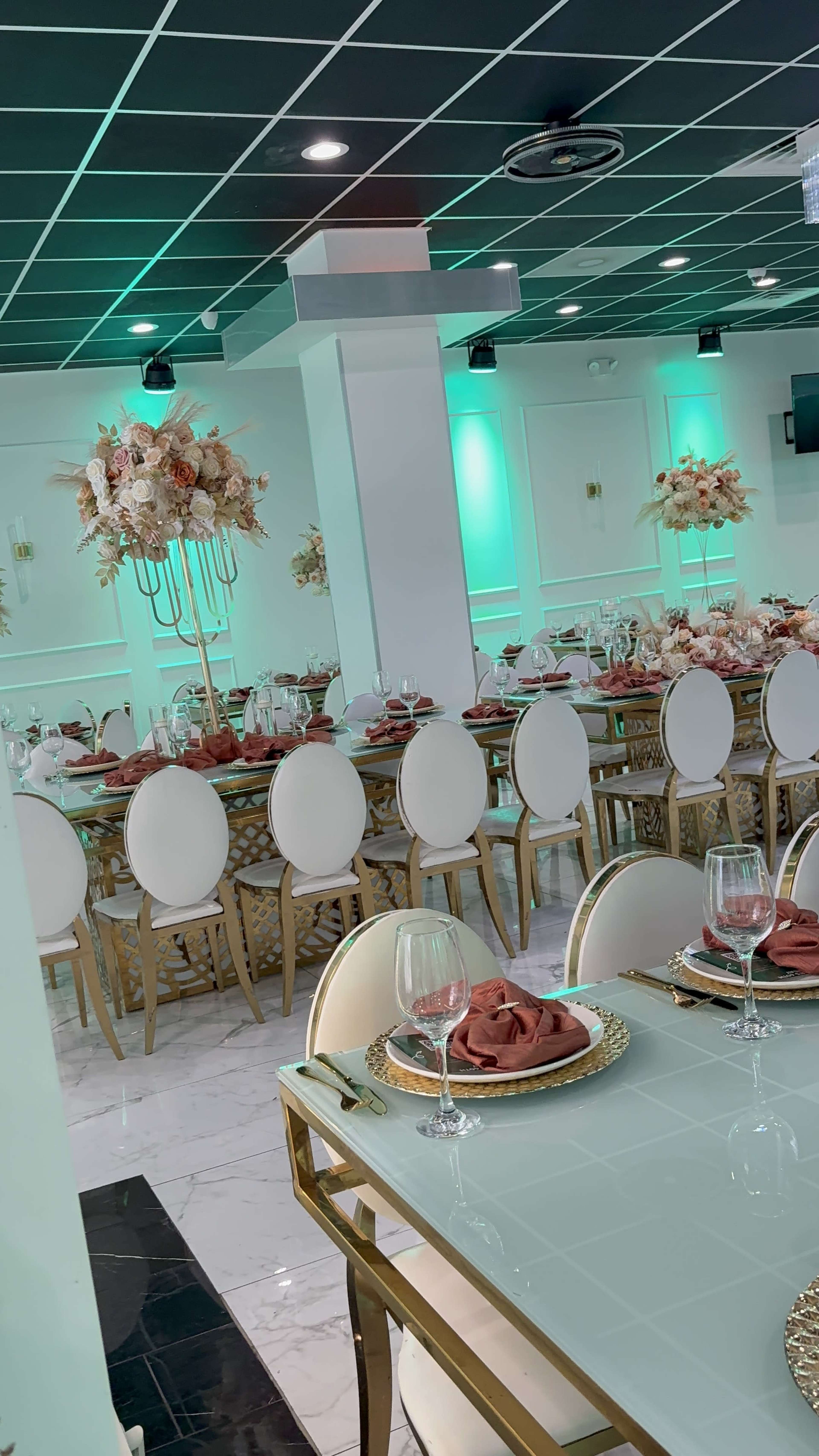 The image shows a banquet hall set for a formal event, featuring elegantly arranged tables dressed with white chairs, gold accents, and floral centerpieces under green lighting.