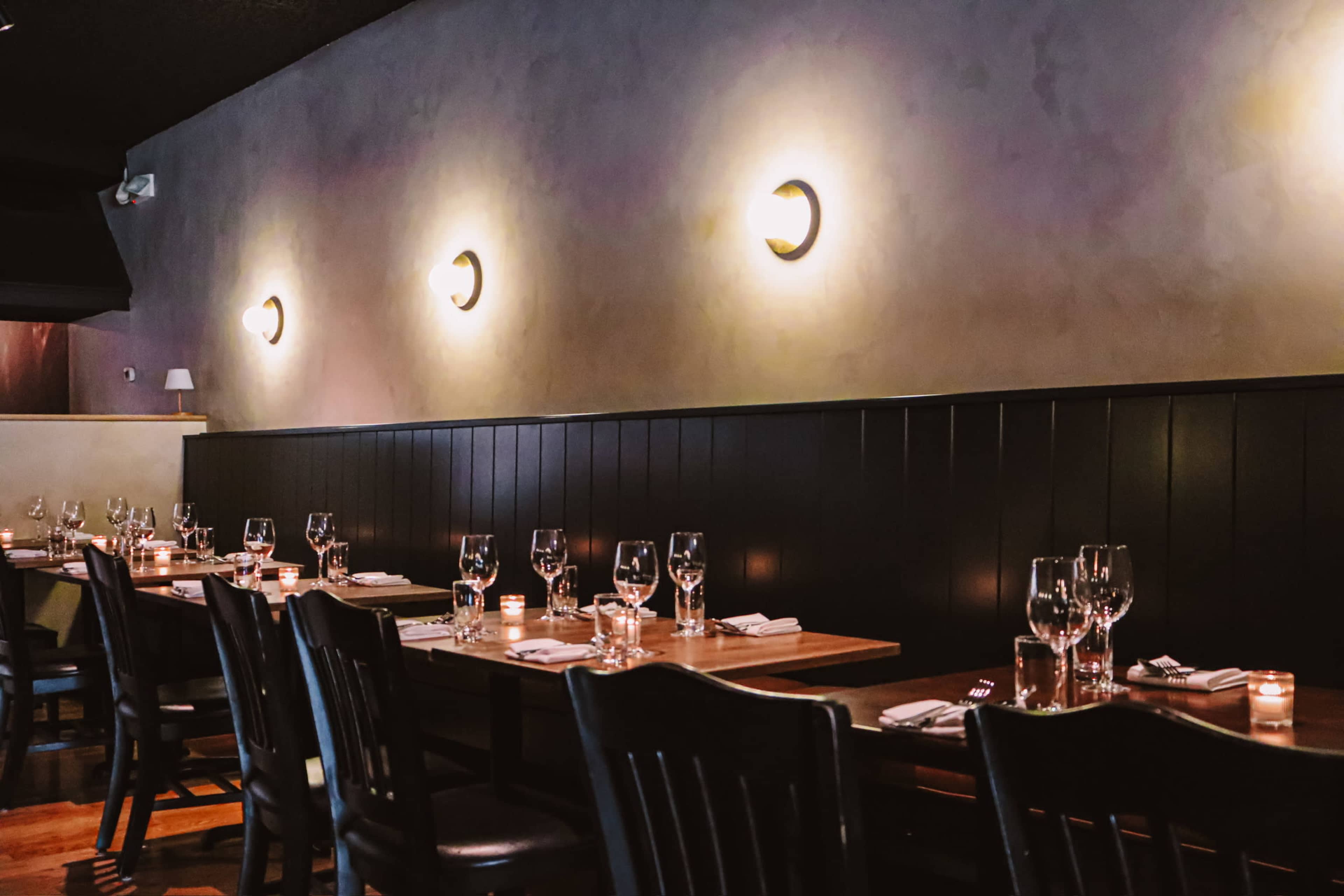 The image shows a dimly lit restaurant interior with wooden tables set with glasses and candles along a black-paneled wall adorned with wall sconces.