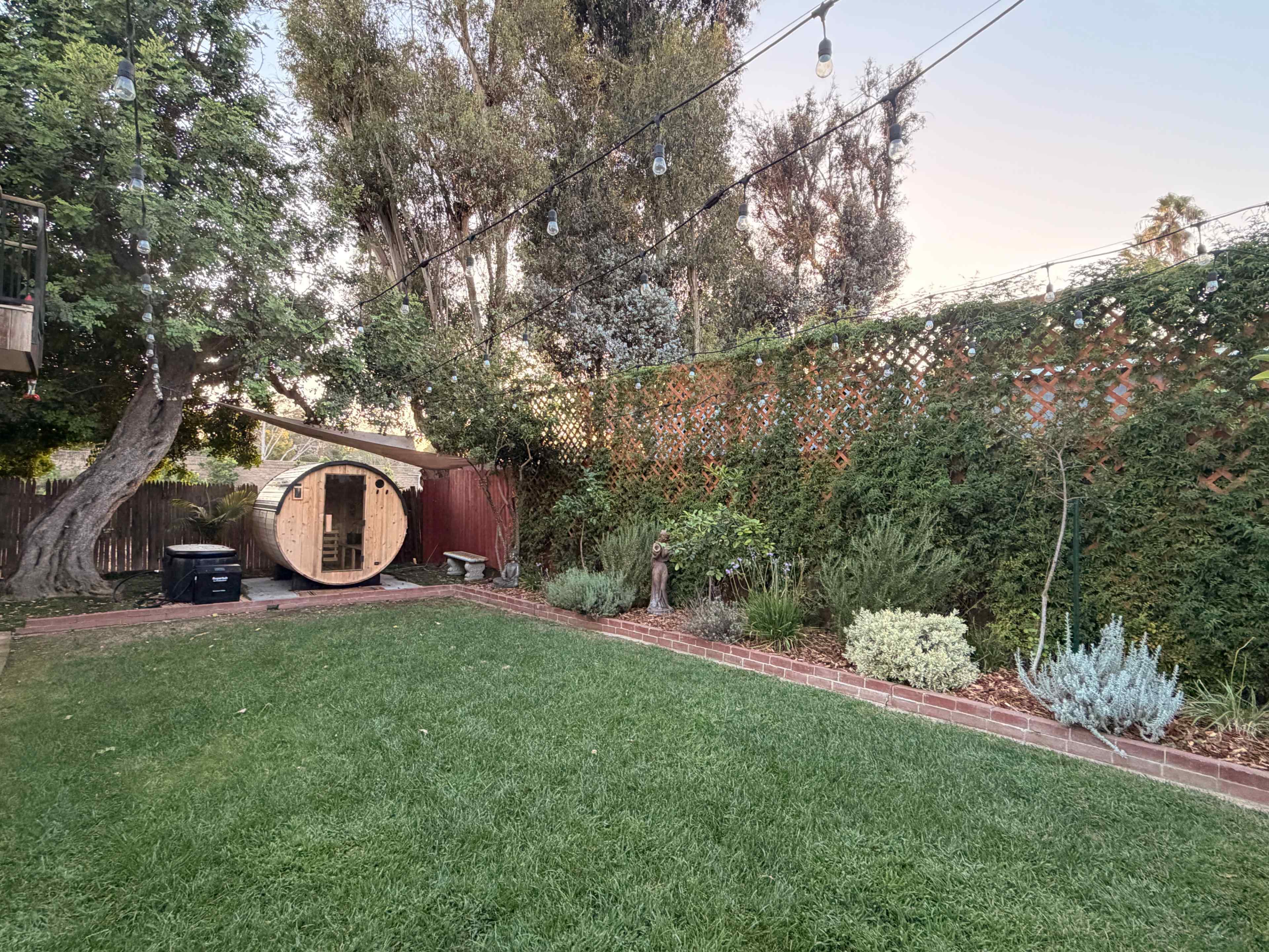 The image shows a backyard with a well-maintained lawn, a tree, a round wooden structure, and various shrubs and plants along the fence.
