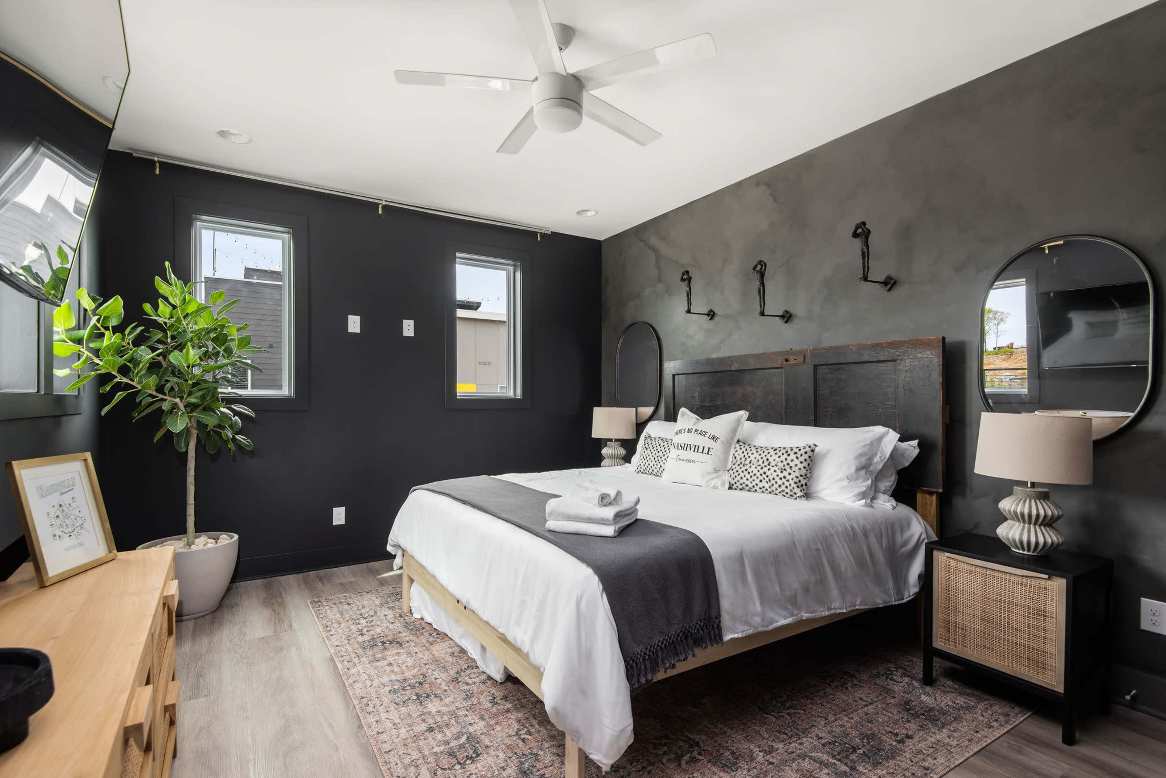 A modern bedroom featuring a double bed with white linens, a gray throw, and a fan overhead, surrounded by dark walls, windows, and decorative elements like a plant and lamps.
