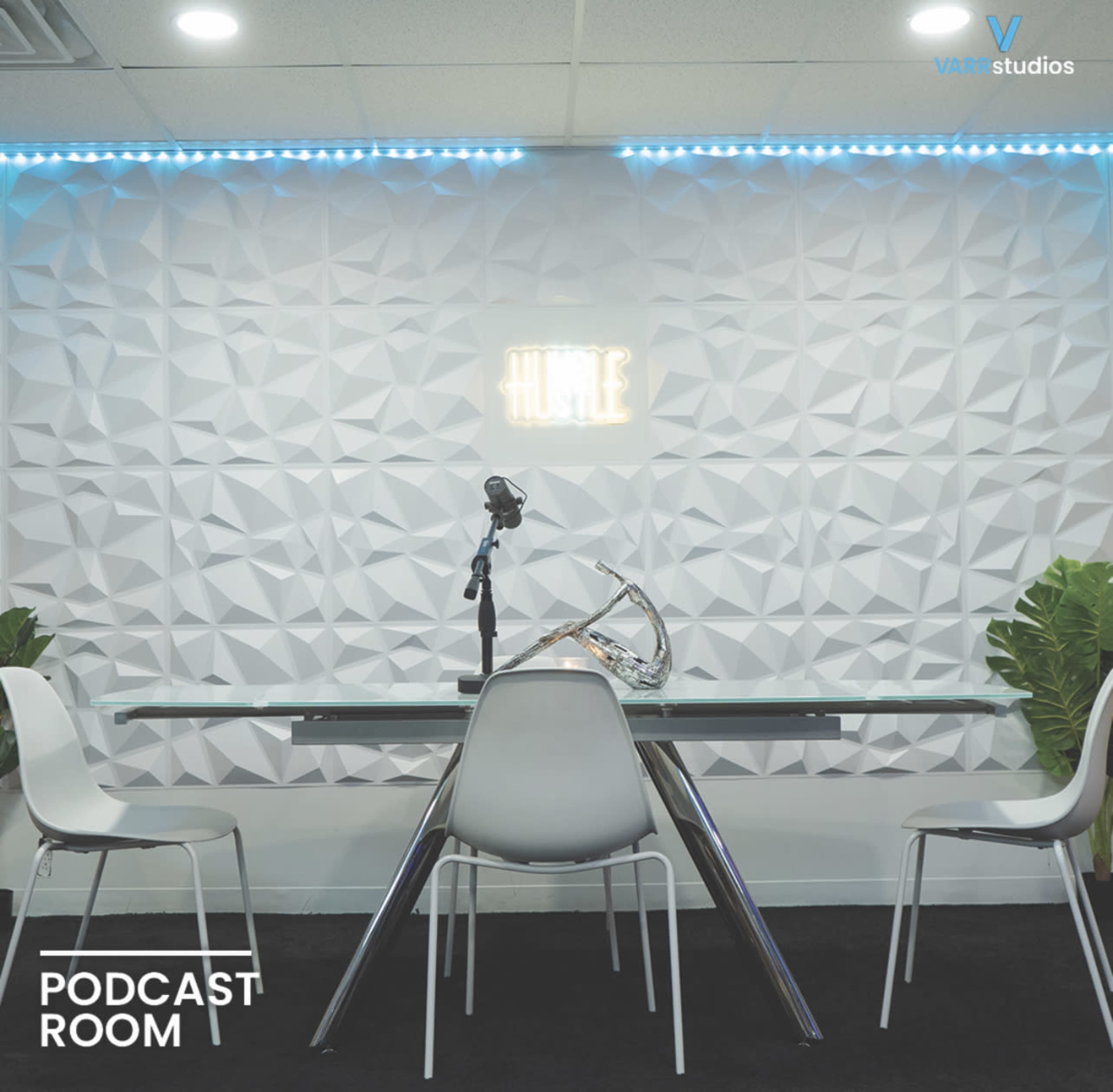 A modern podcast room features a glass table, white chairs, a microphone, and a textured wall with a neon sign that reads "HUSTLE."