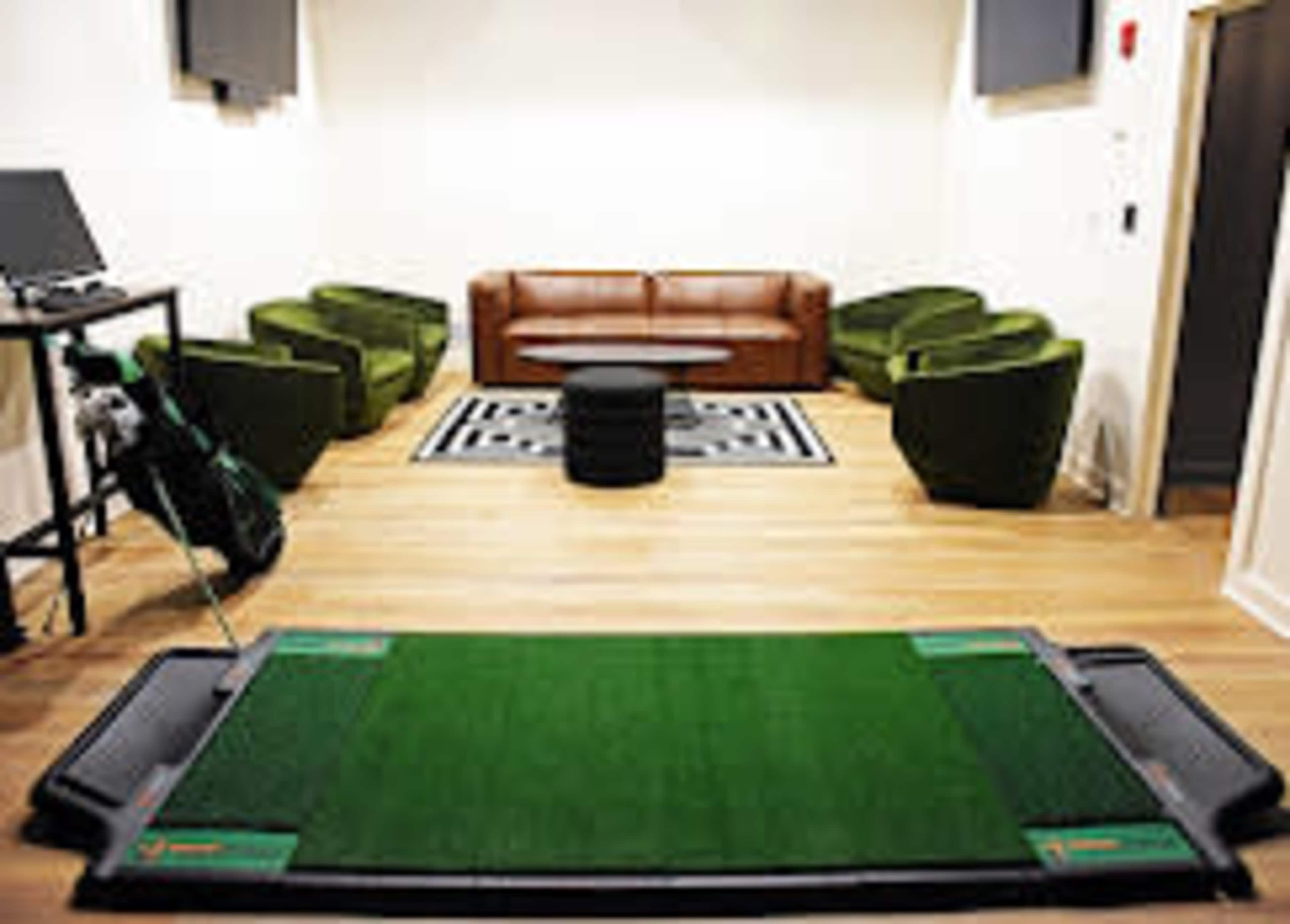 The image shows a room with a green golf practice mat in the foreground, a brown leather sofa, and several green chairs arranged around a black coffee table.