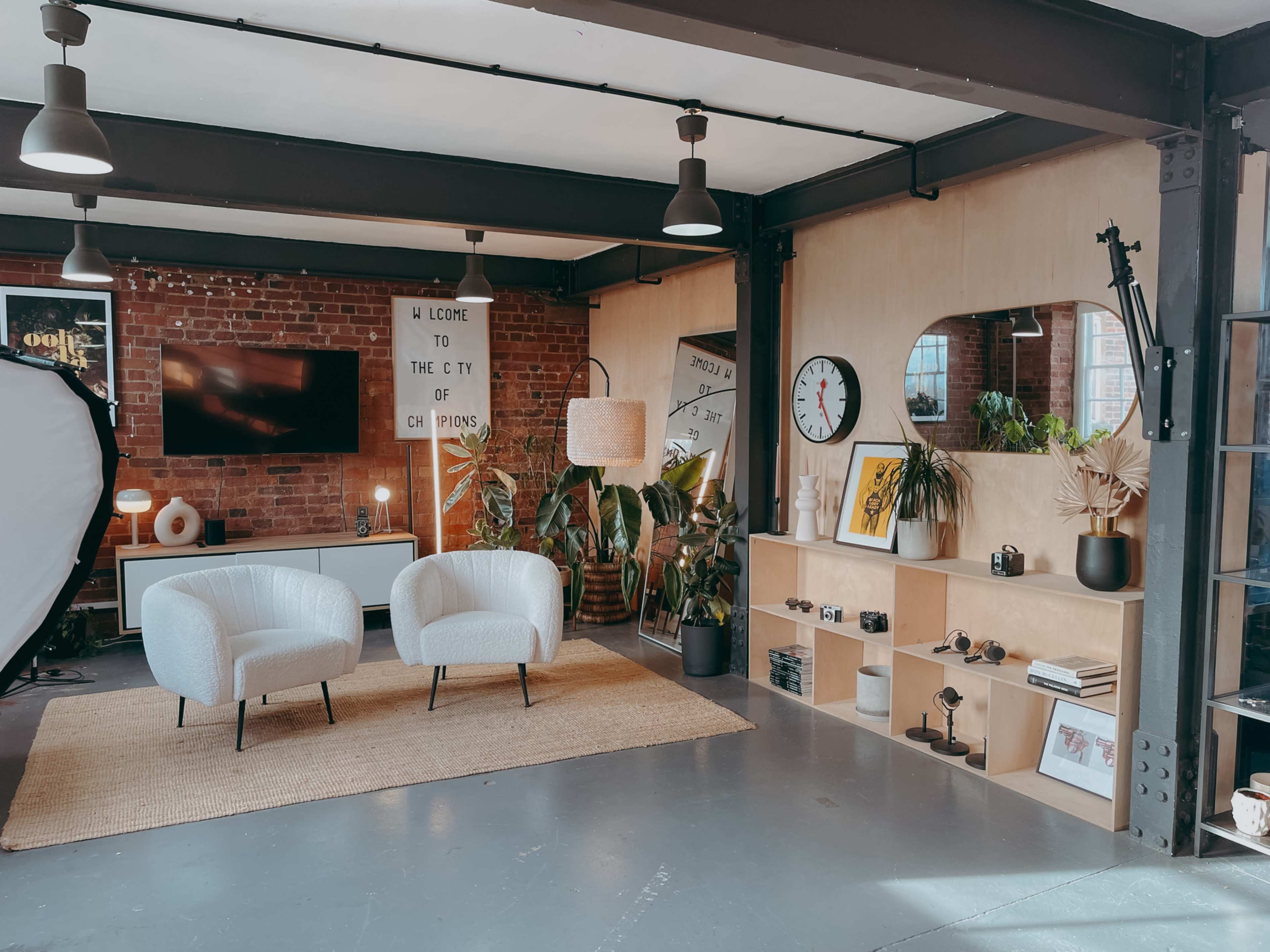 The image shows a modern interior space featuring two white armchairs, a large wall mirror, a wall-mounted clock, various plants, and an organized shelf displaying decorative items and a television.