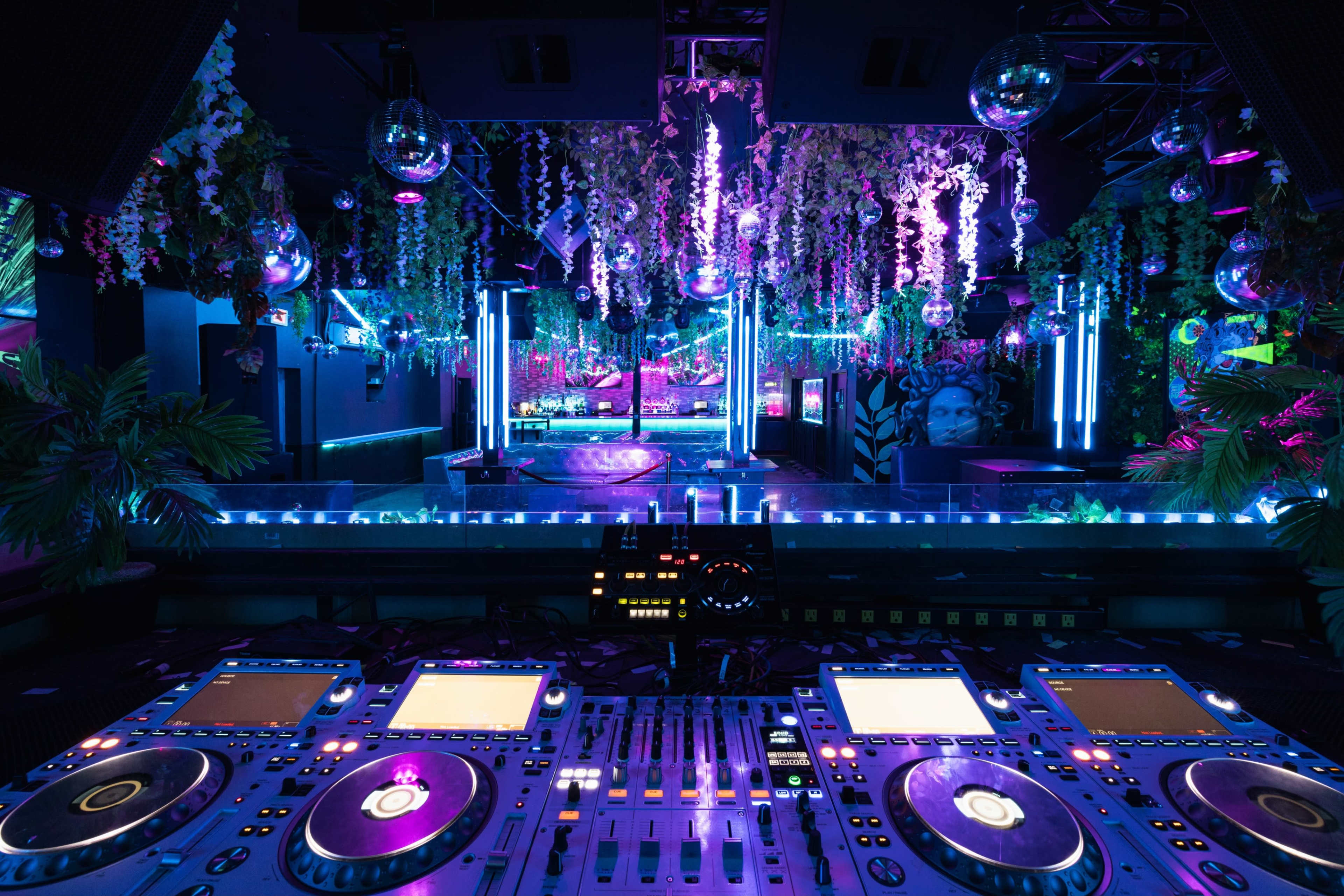 A DJ booth with turntables and mixers is located at the foreground, surrounded by vibrant neon lights and decorative plants in a lively nightclub setting.