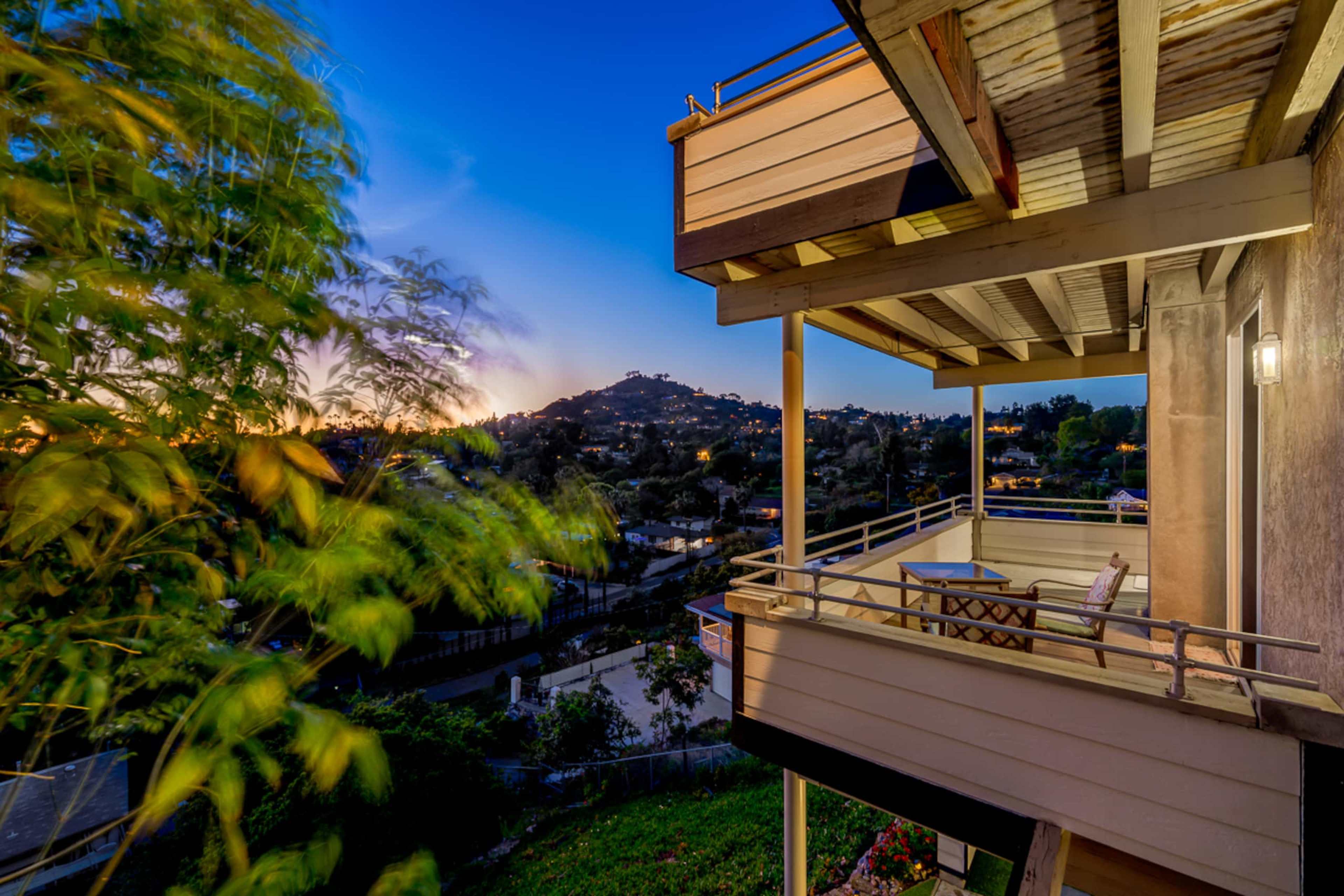 The image shows a balcony overlooking a hillside neighborhood during twilight, with a vibrant sky and greenery swaying in the foreground.