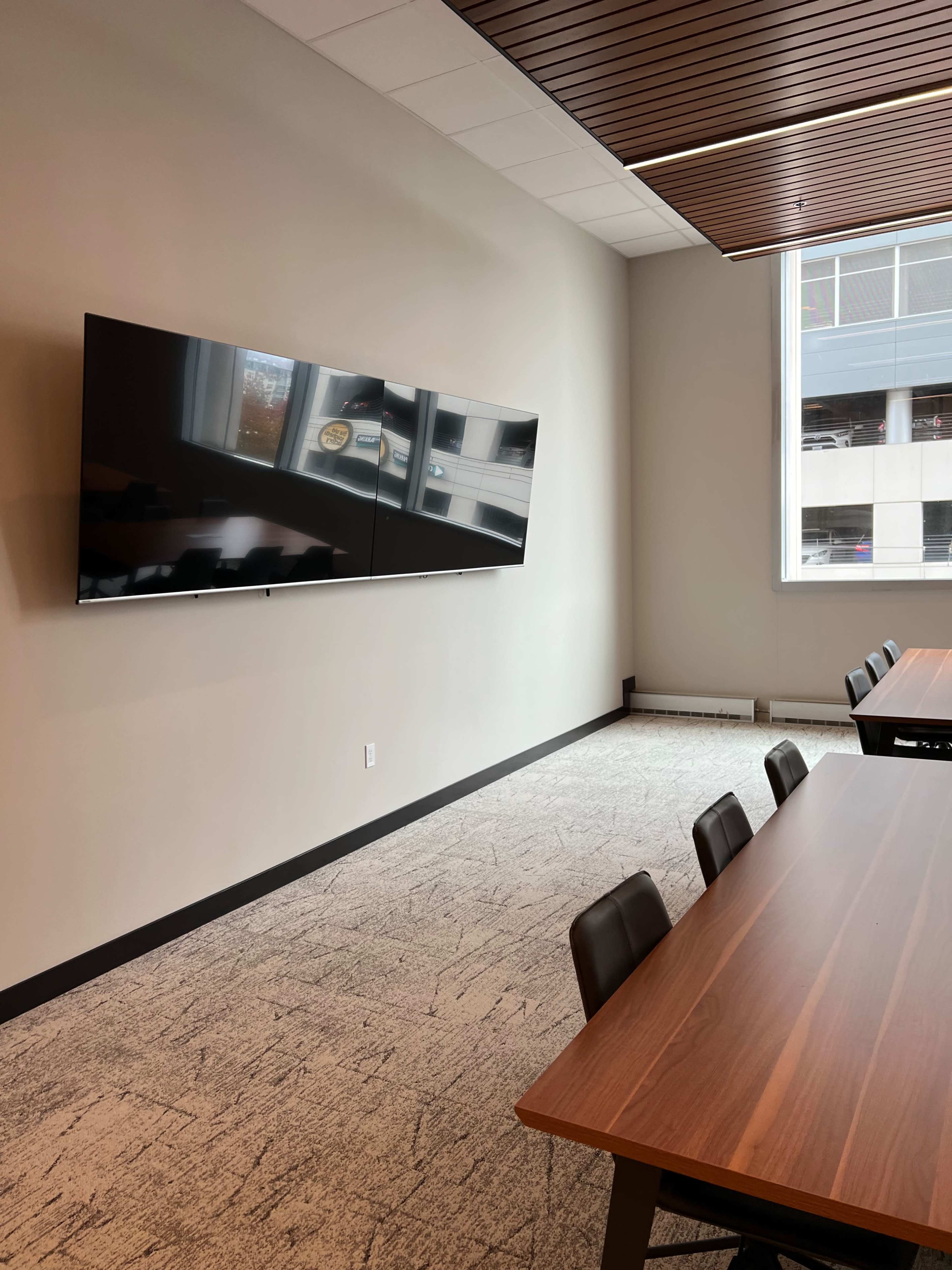 A modern conference room features a large flat-screen display mounted on the wall opposite a long wooden table with black chairs.