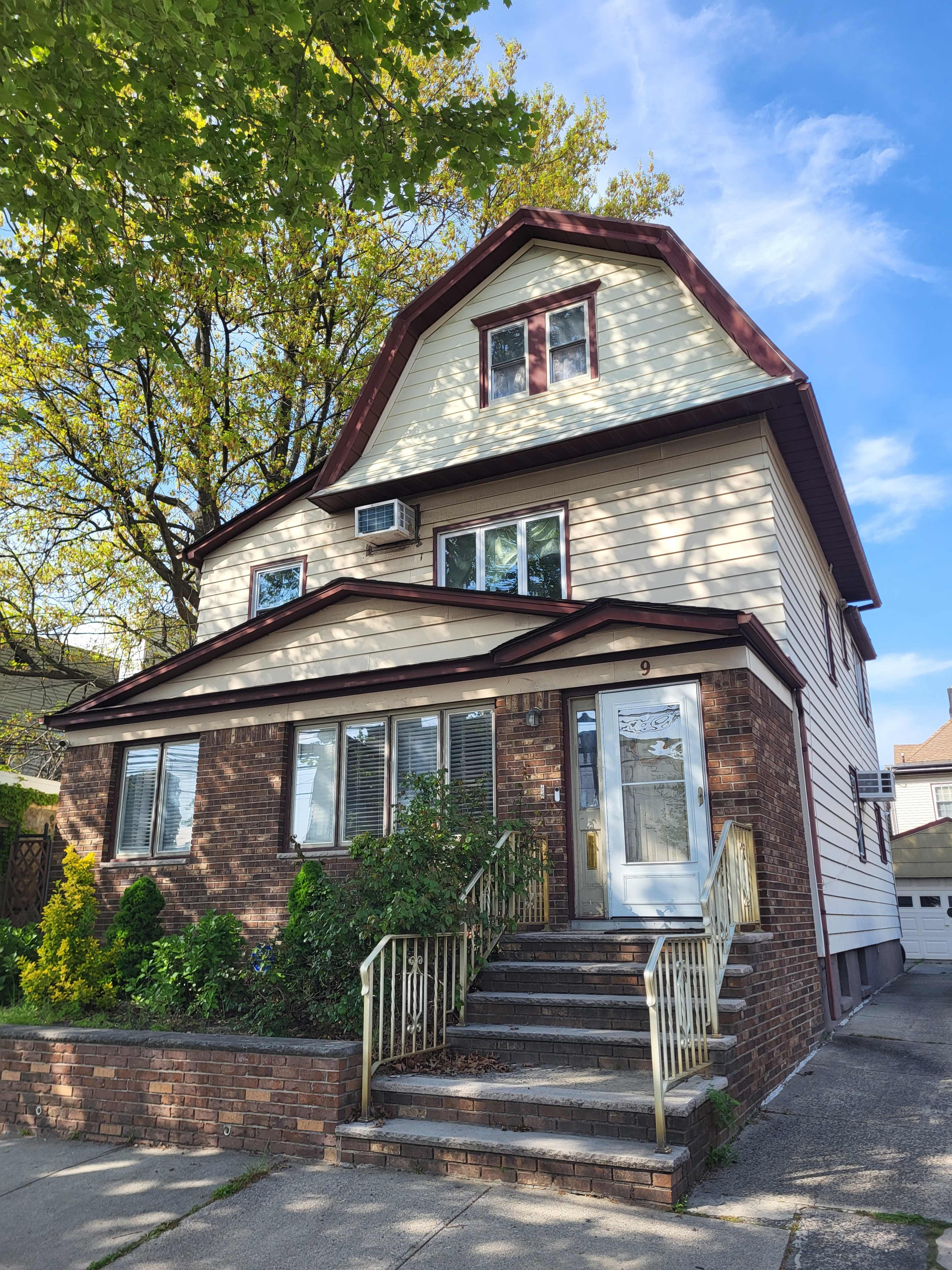 A three-story house with a gabled roof features a mix of brick and siding exterior, an air conditioning unit, and a front staircase with a railing.