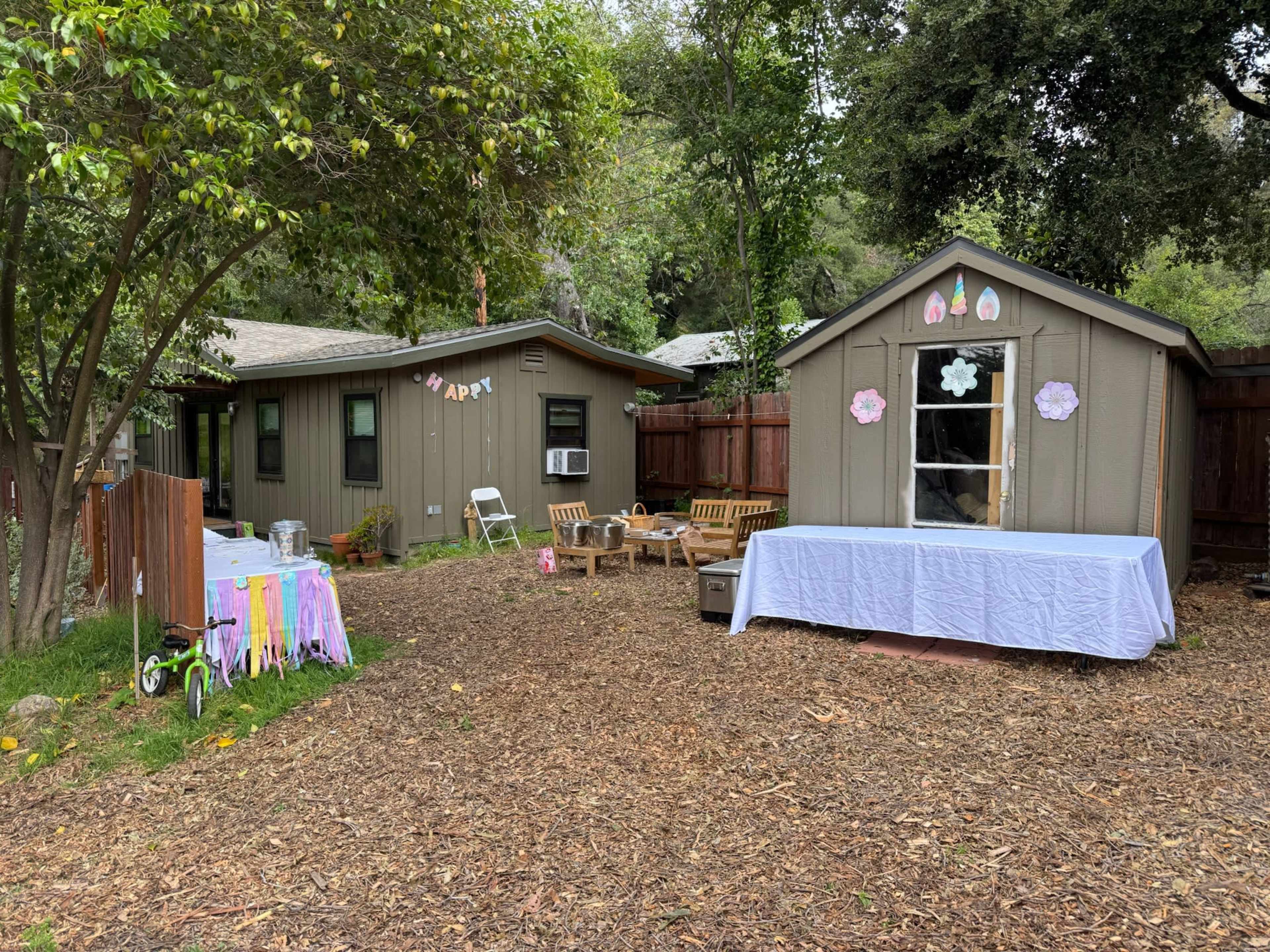 The image shows a backyard area featuring a small cabin, a shed decorated with flowers, outdoor seating, and a table covered with a white tablecloth.