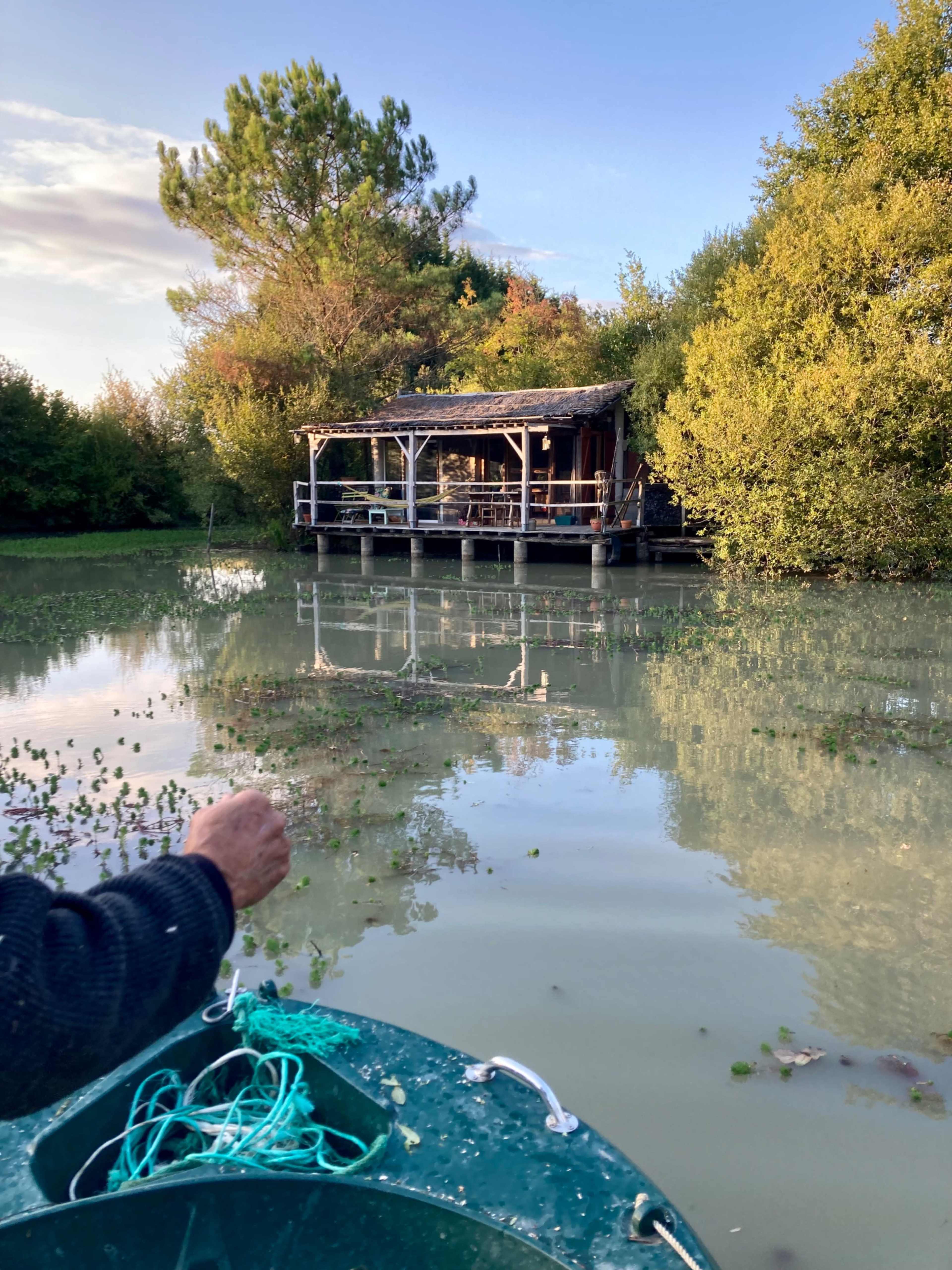 A person holds a fishing line while looking at a wooden cabin surrounded by trees, reflecting in the calm water.