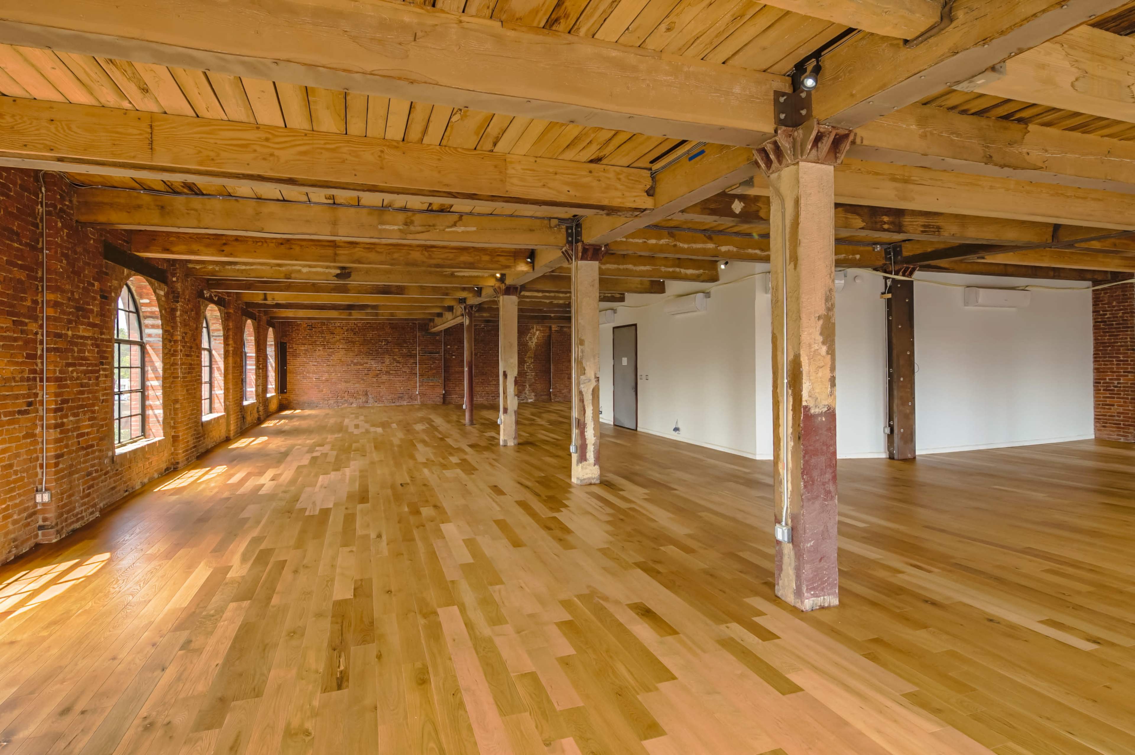 The image shows a spacious, open room with wooden flooring, exposed brick walls, and wooden beams overhead.