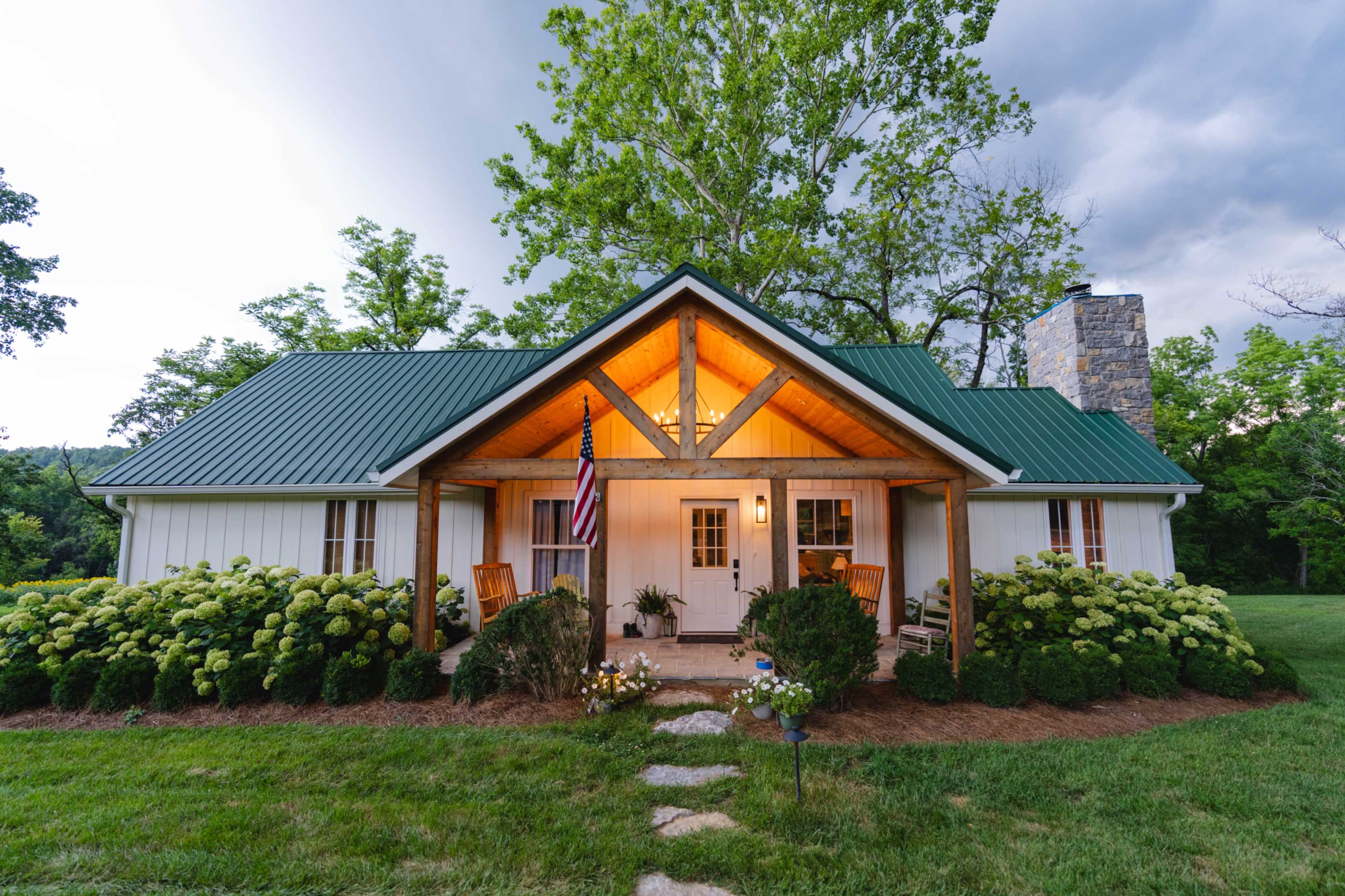 A white cottage with a green metal roof features a front porch adorned with rocking chairs and surrounded by lush greenery and blooming shrubs.