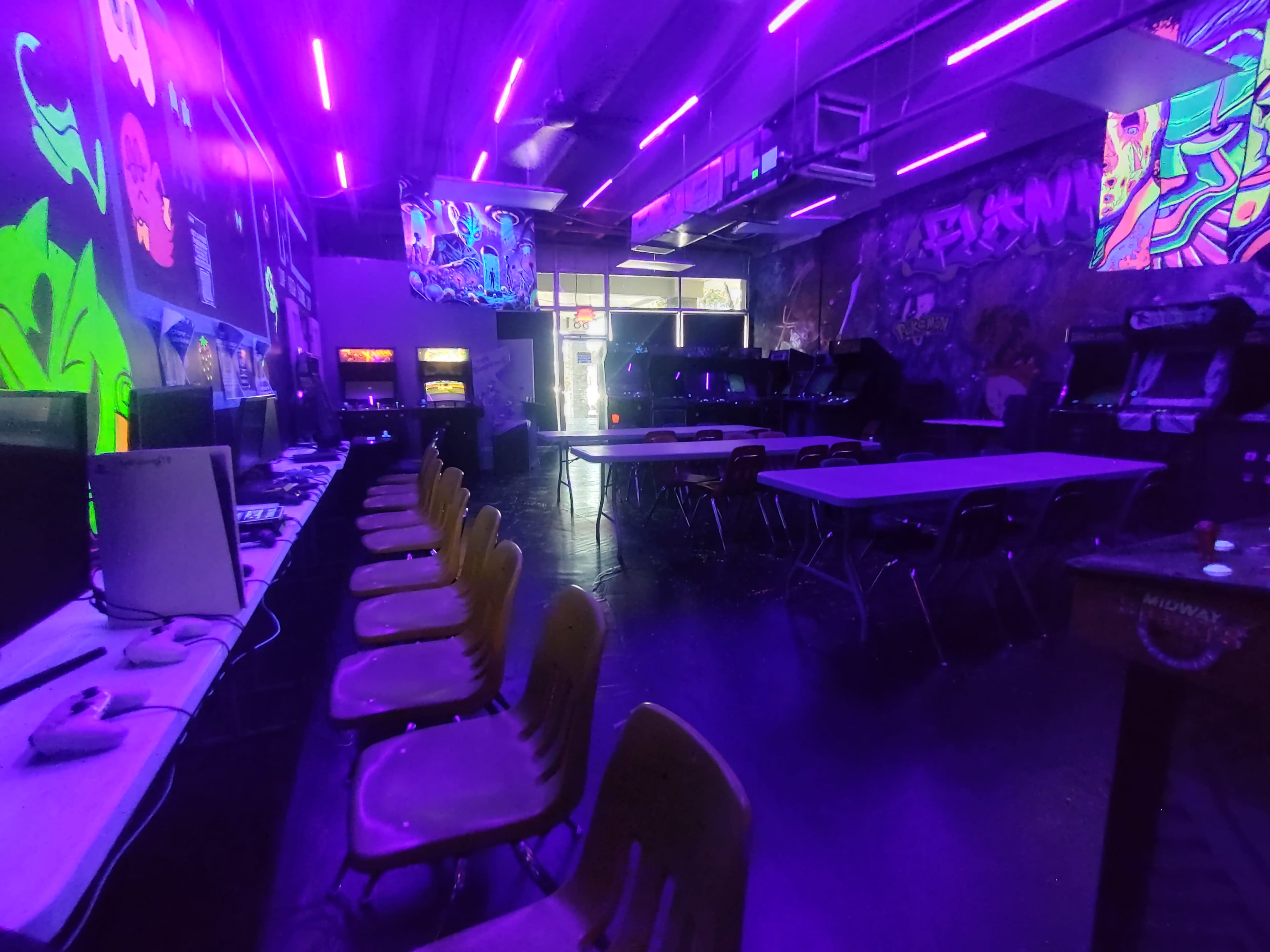 A dimly lit arcade features rows of chairs and tables under purple neon lighting, with colorful wall art and vintage arcade machines in the background.