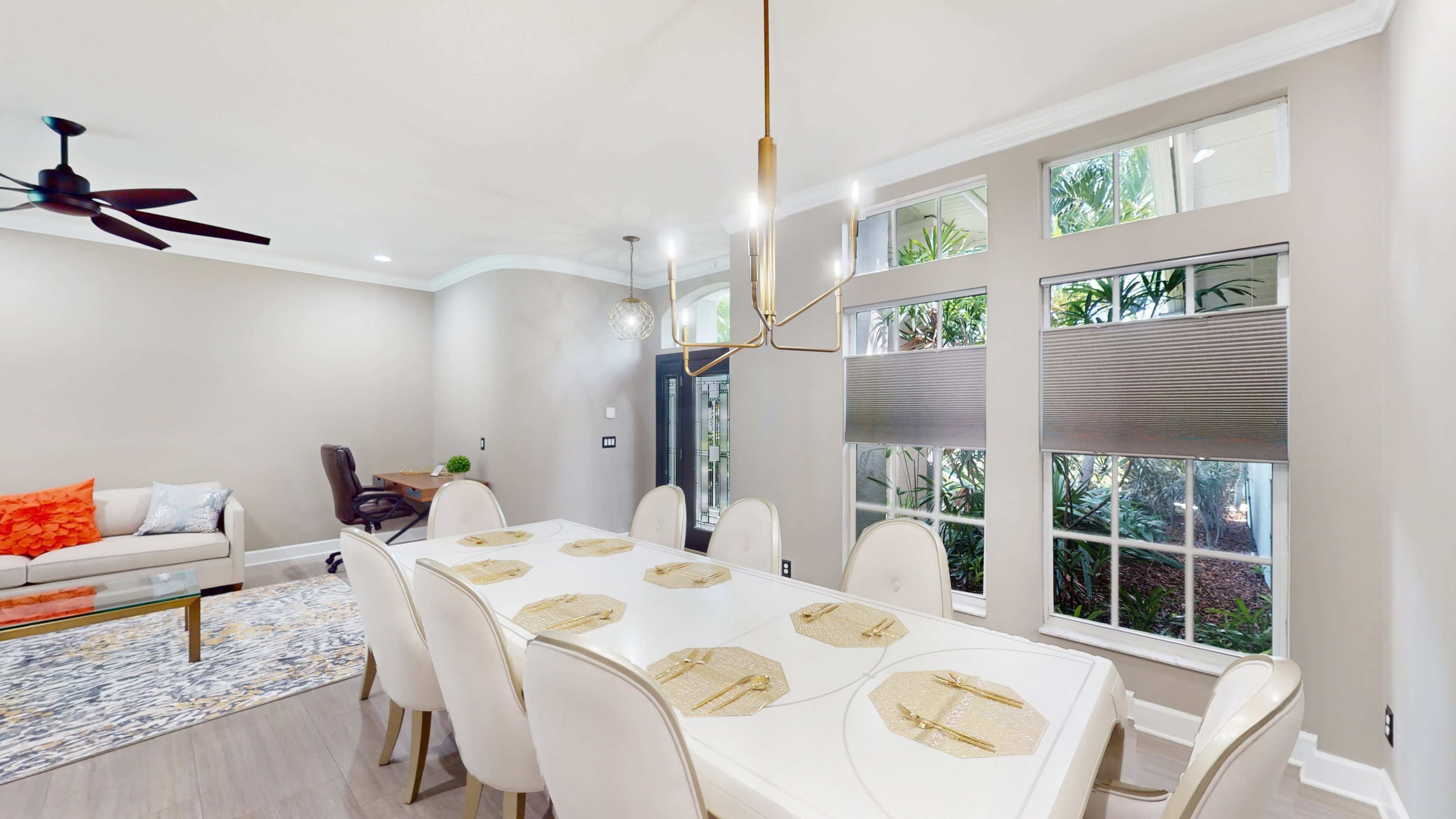 A modern dining area features a long white table with beige chairs, surrounded by large windows that provide natural light and views of greenery.