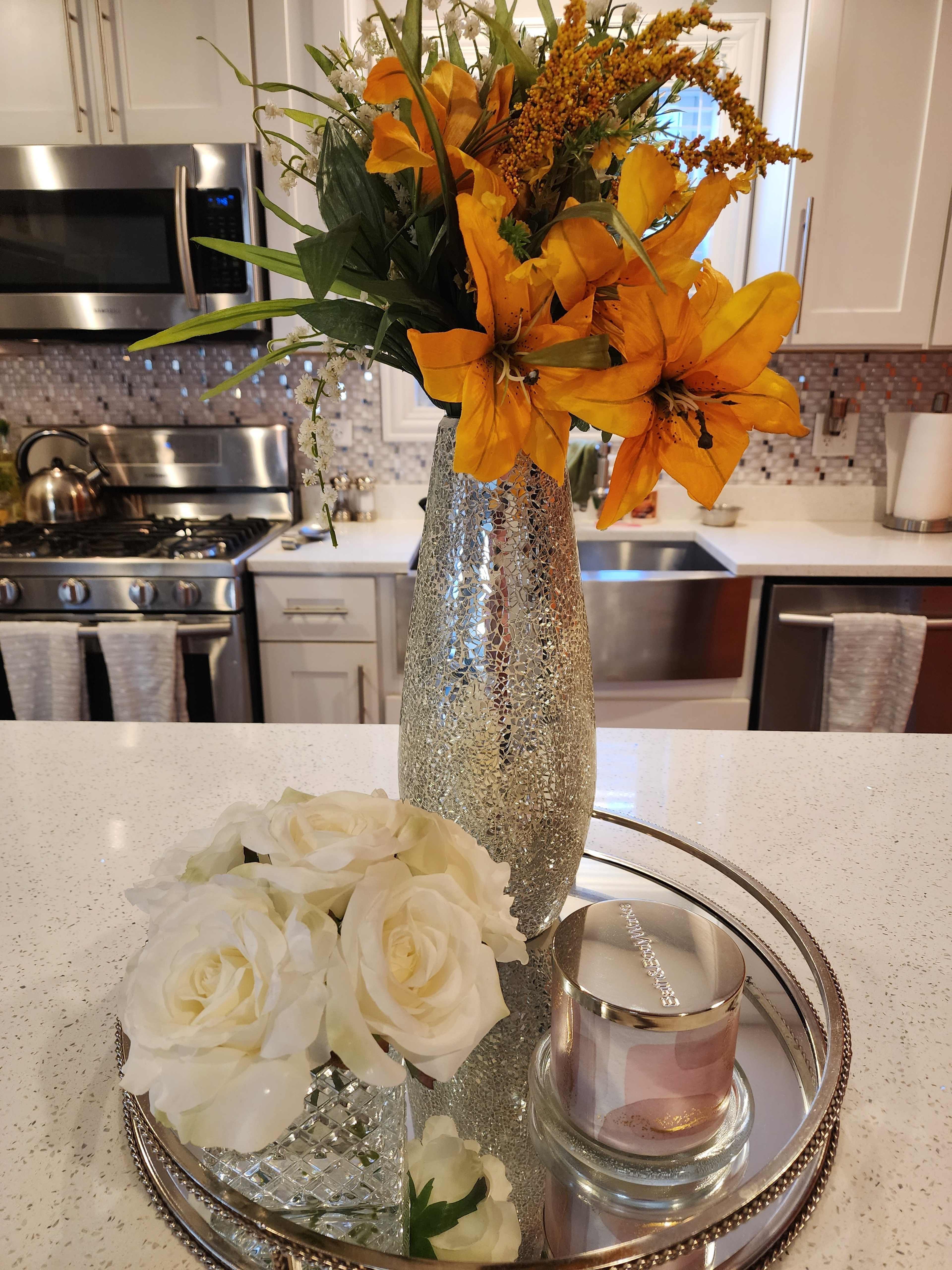 A vase of orange flowers stands beside a small arrangement of white roses and a candle on a mirrored tray on a kitchen countertop.