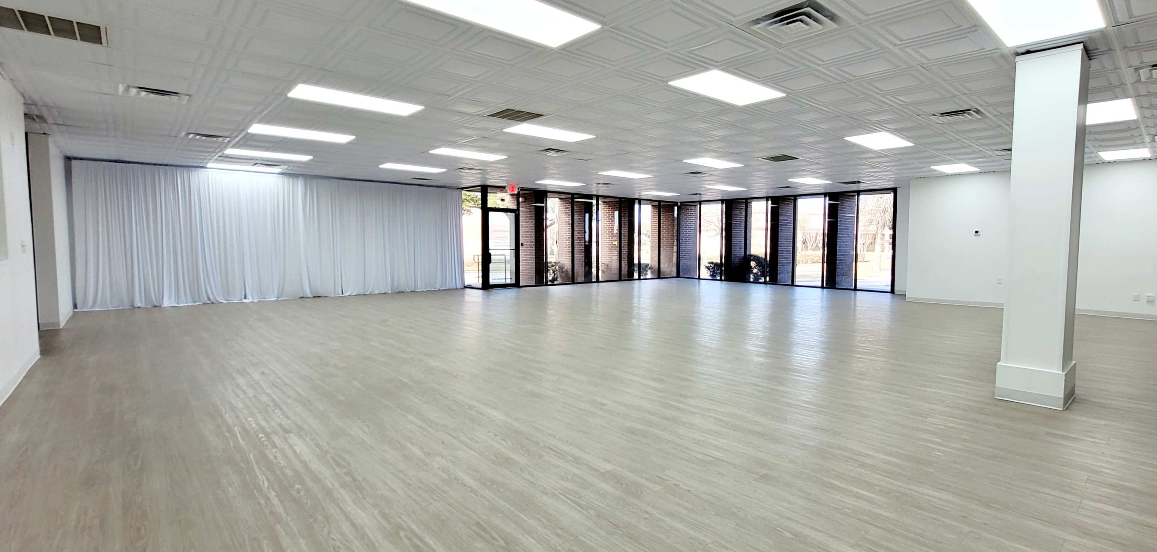 The image shows a spacious, empty interior room with a light wood floor, large windows, and white walls.