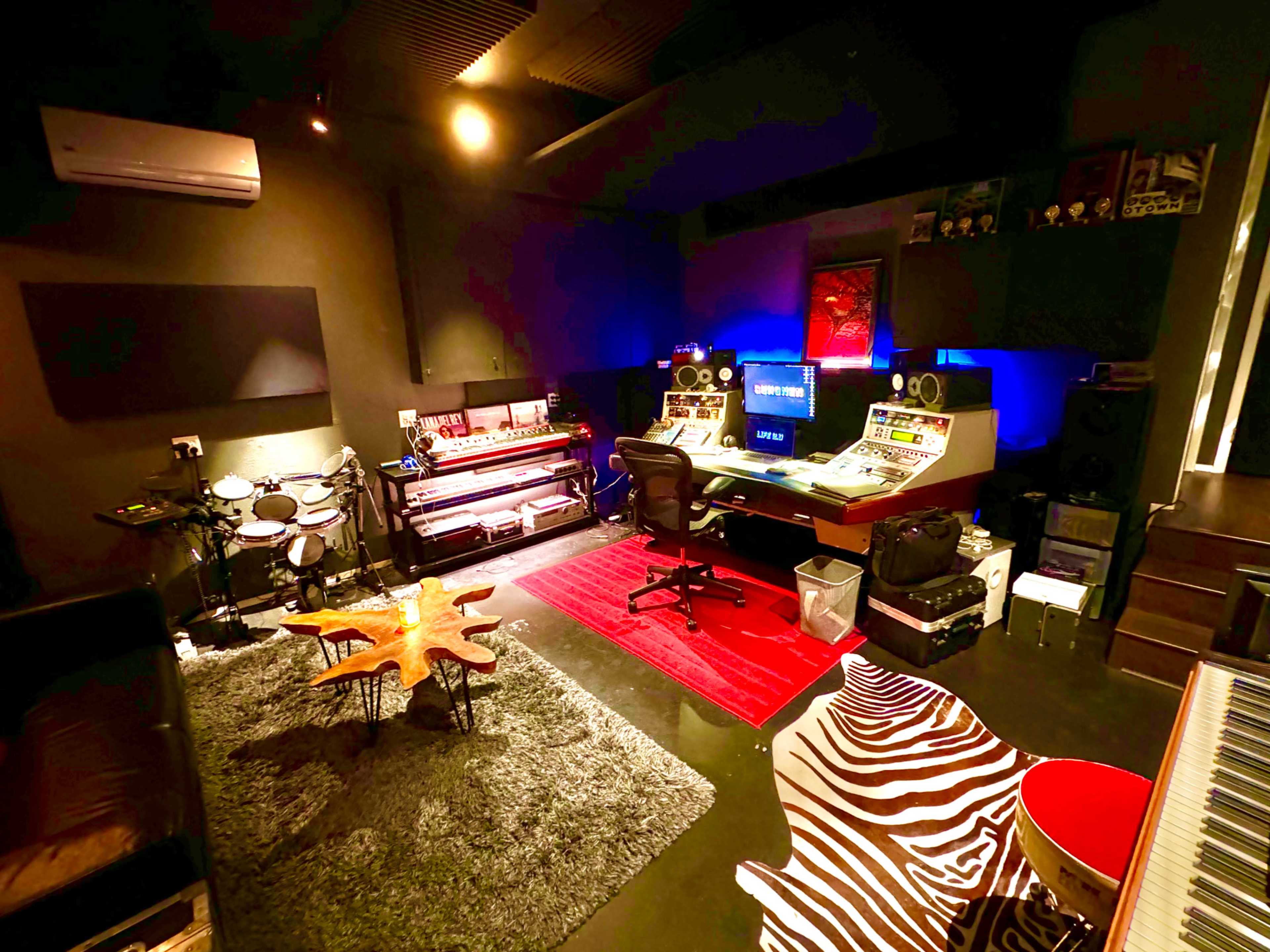The image shows a music production studio featuring a console with monitors, an electric piano, electronic drum set, and area rugs on the floor.