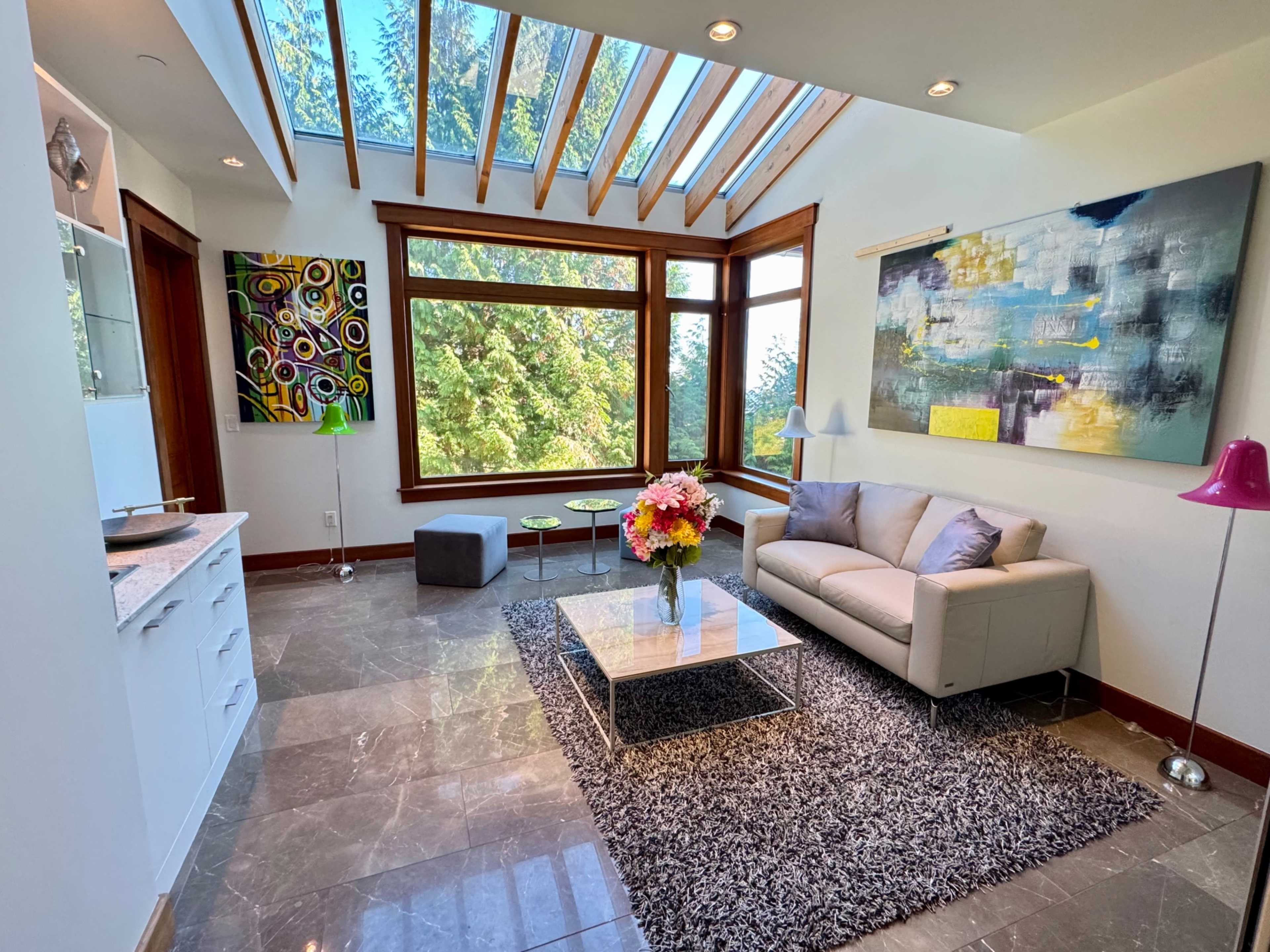 The image shows a bright, modern living room with large windows, a plush sofa, a glass coffee table, and contemporary artwork on the walls.
