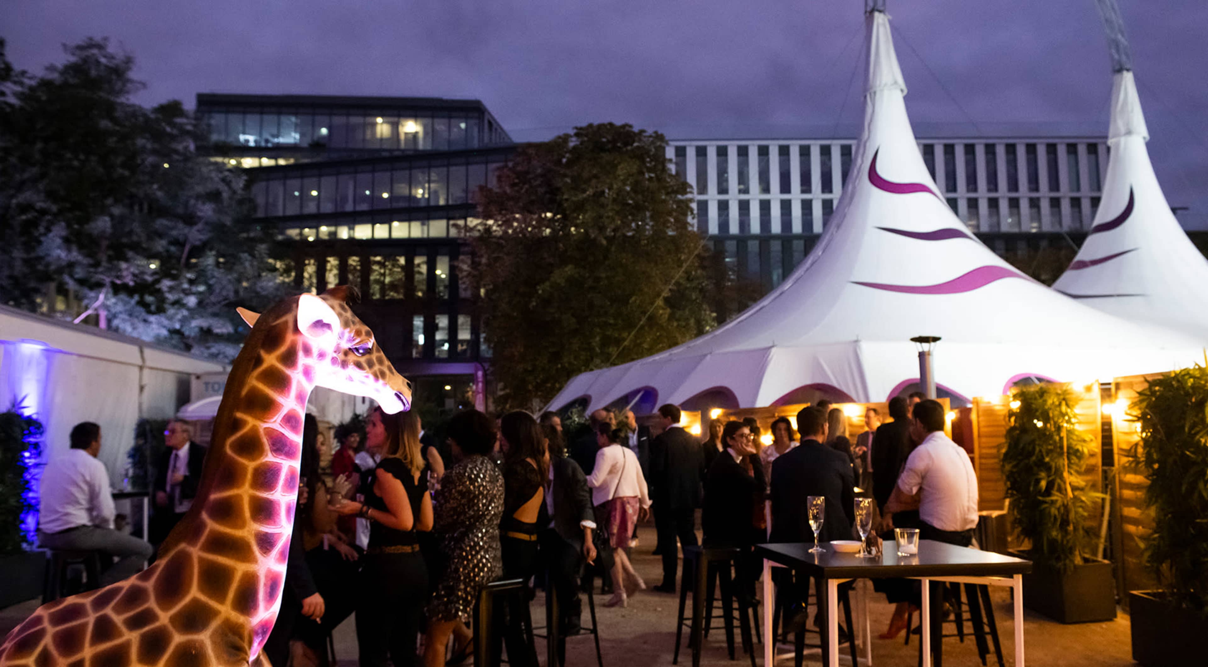 A giraffe sculpture with neon lighting stands in the foreground of a festive outdoor event featuring a large tent and groups of people socializing.