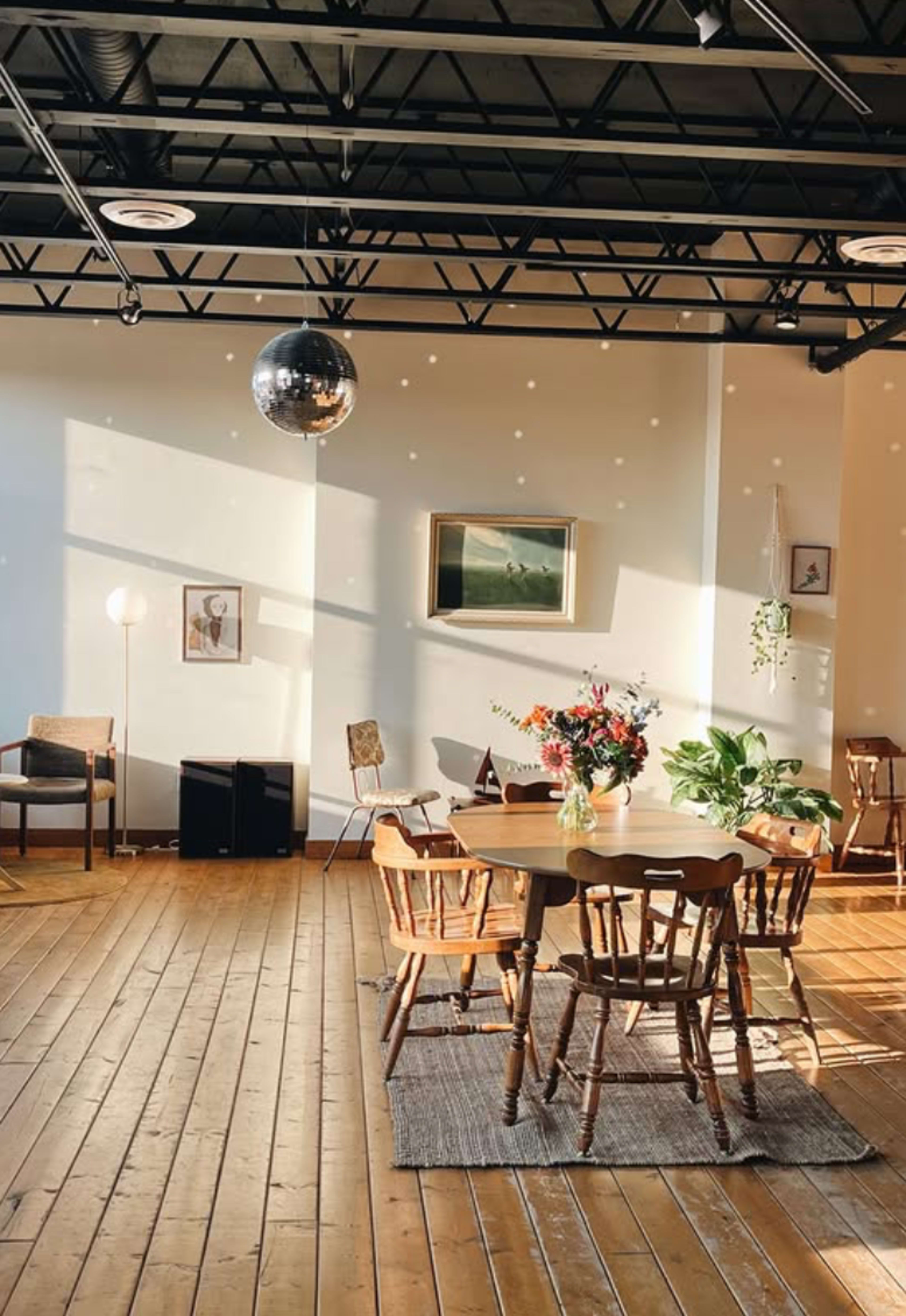 The image shows a spacious room with wooden flooring, a round table surrounded by chairs, a large disco ball hanging from the ceiling, and potted plants near the window.