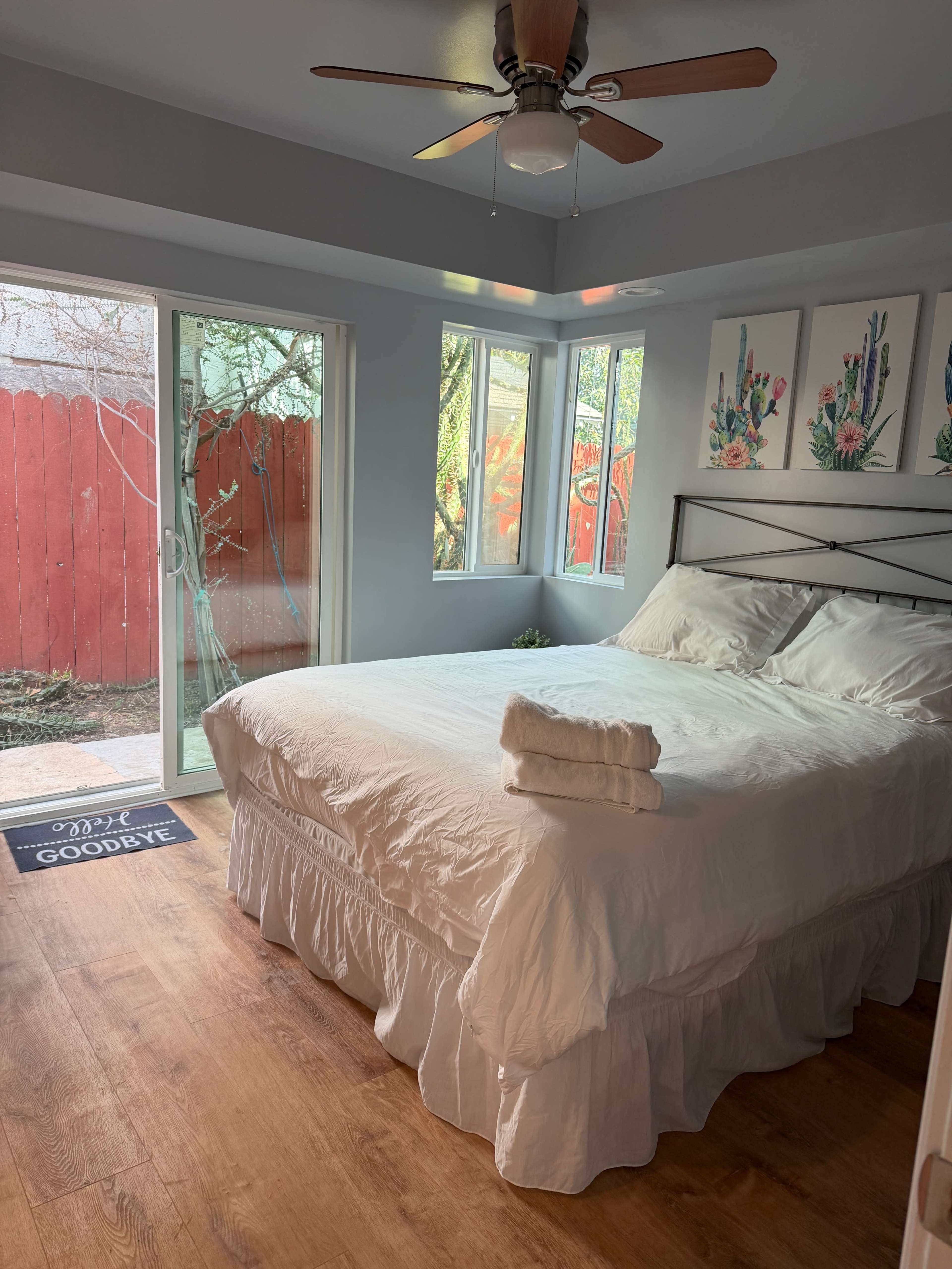 The image shows a bright bedroom with a white bedspread, framed artwork on the walls, and glass doors leading to a small outdoor area.