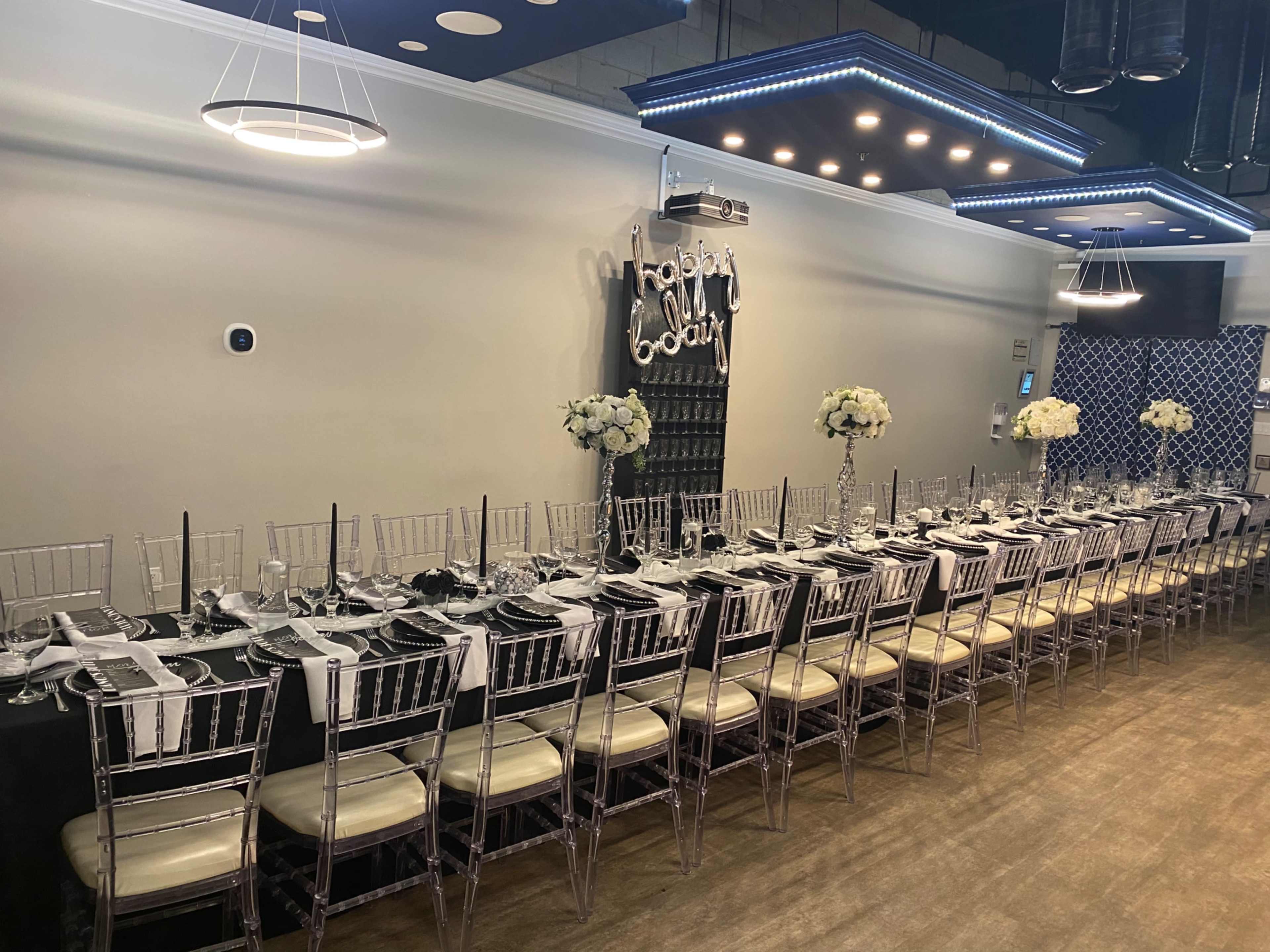 A long banquet table is set up with white tableware and black accents, flanked by transparent chairs, in a decorated dining space.