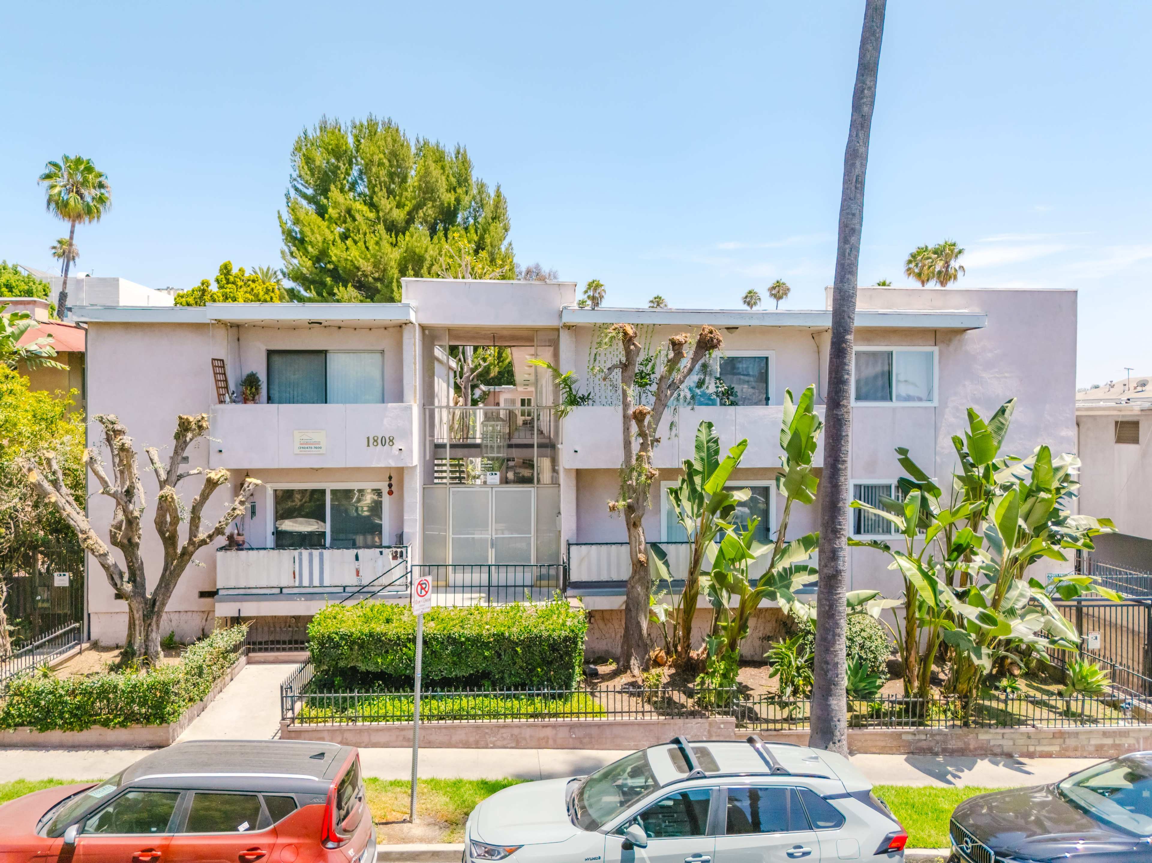 The image shows a two-story apartment building surrounded by palm trees and banana plants, with parked cars lining the street in front.