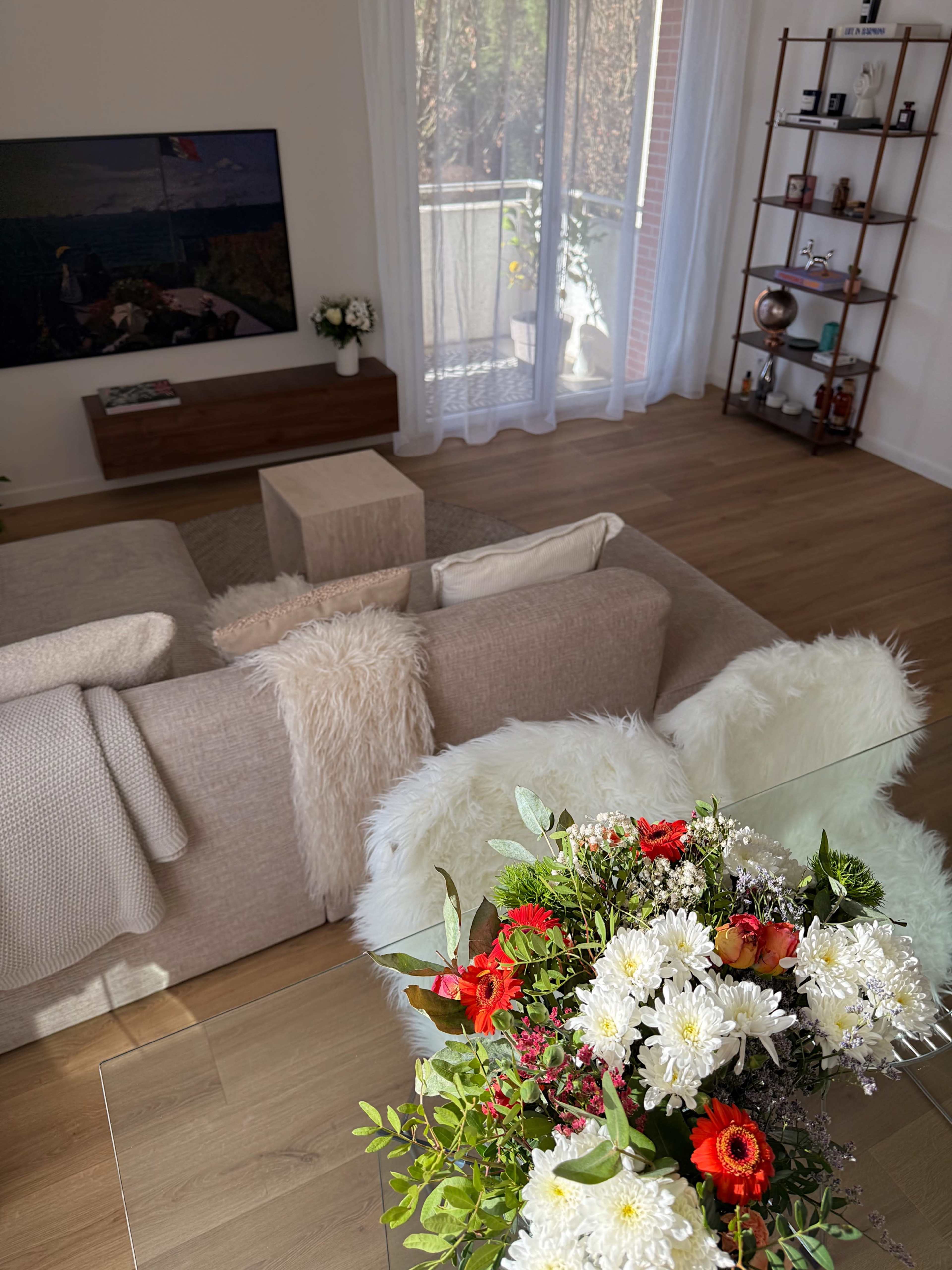 The image shows a well-lit living room with a beige sofa, a wooden coffee table, and a decorative shelf, featuring a vase of colorful flowers in the foreground.