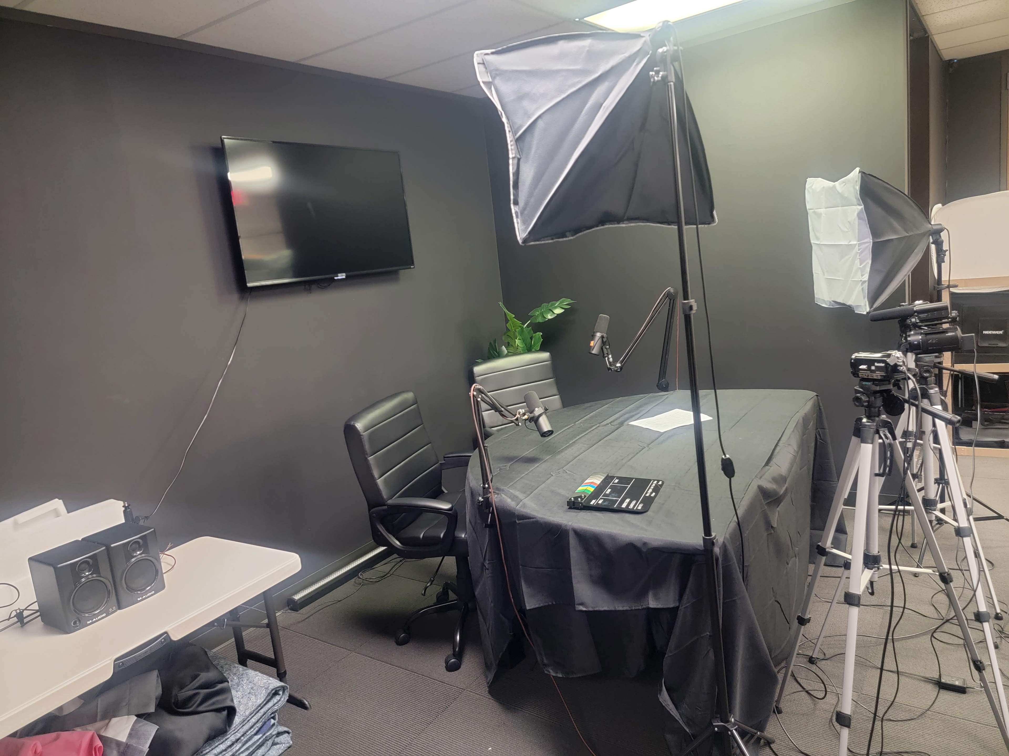 A recording studio setup features a black table, microphones, camera tripods, and a flat-screen TV mounted on a dark wall.