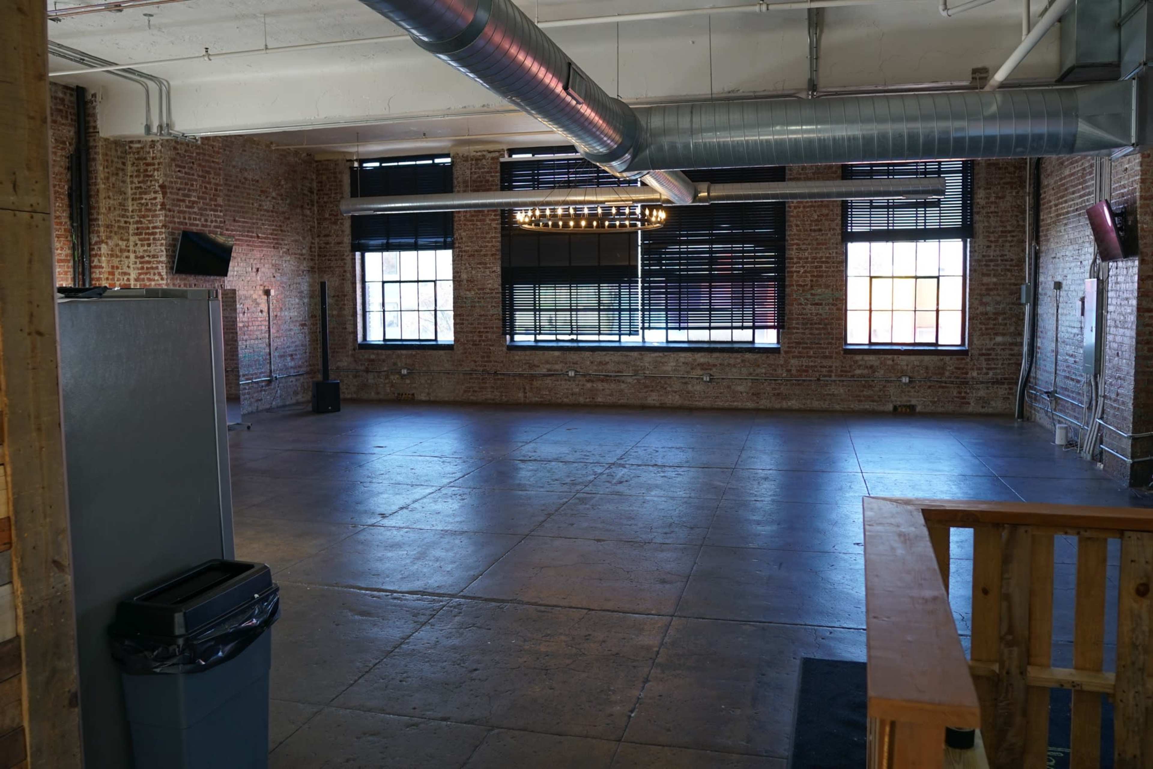 An empty room with exposed brick walls, large windows with blinds, and a concrete floor.