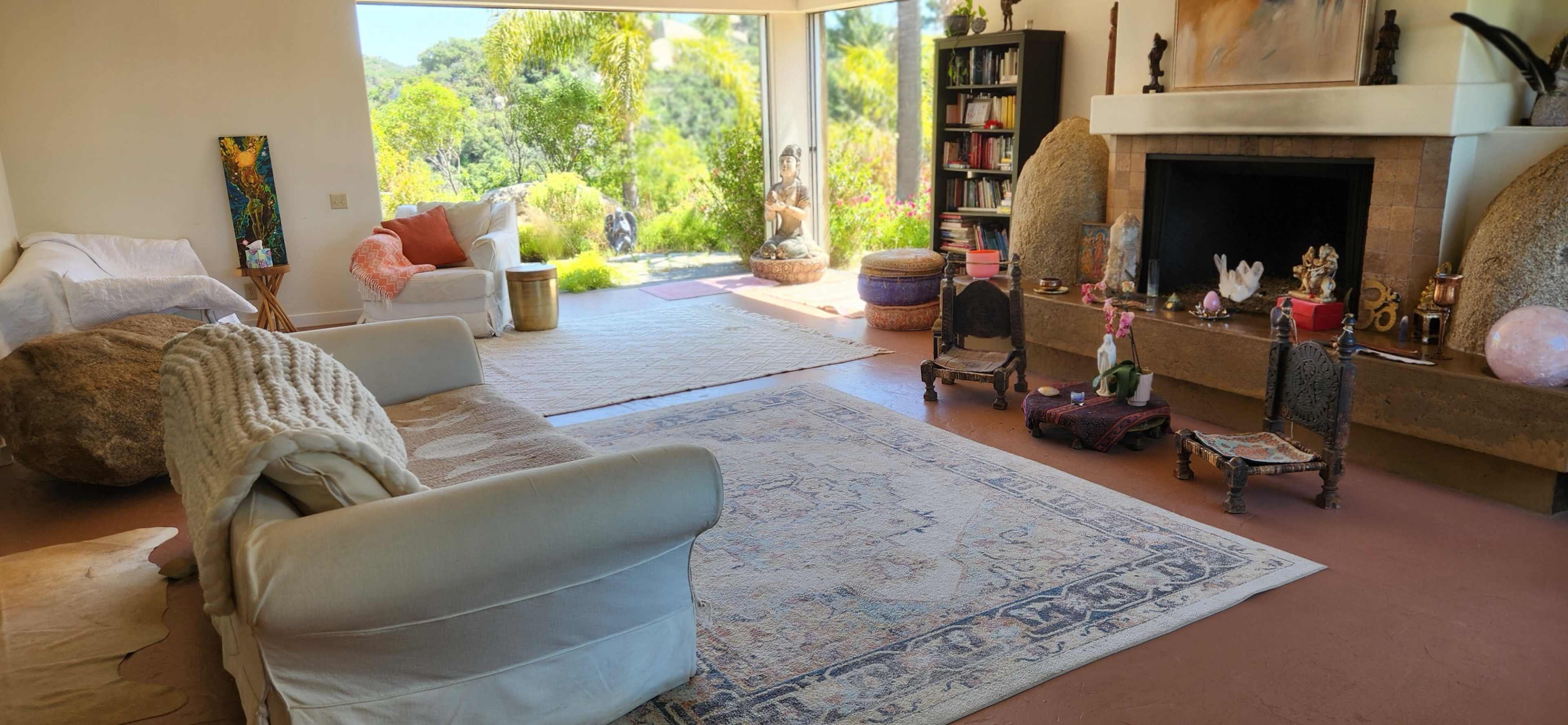 A spacious living room featuring a light-colored sofa, a decorative rug on the floor, and large windows that offer a view of greenery outside.