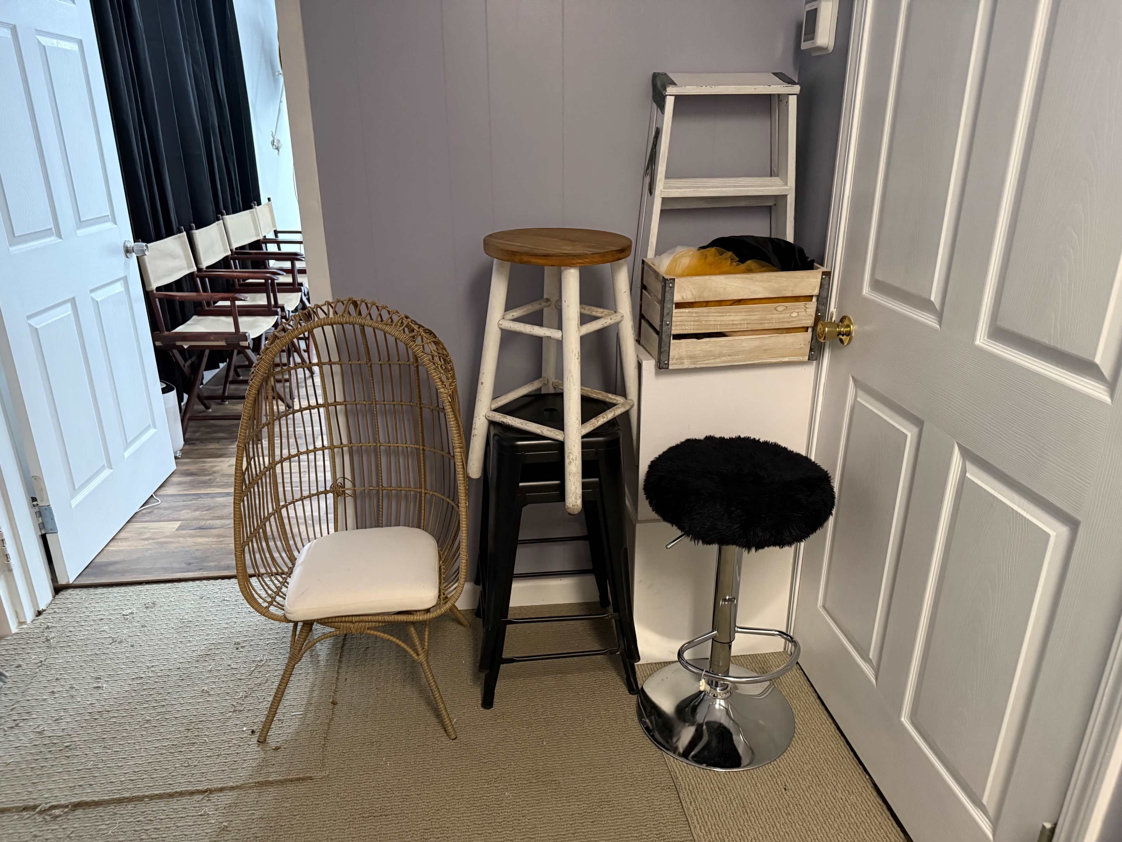 The image shows a small entryway featuring three different stools and a wicker chair against a wall, with a door on the right and a glimpse of additional seating in the background.