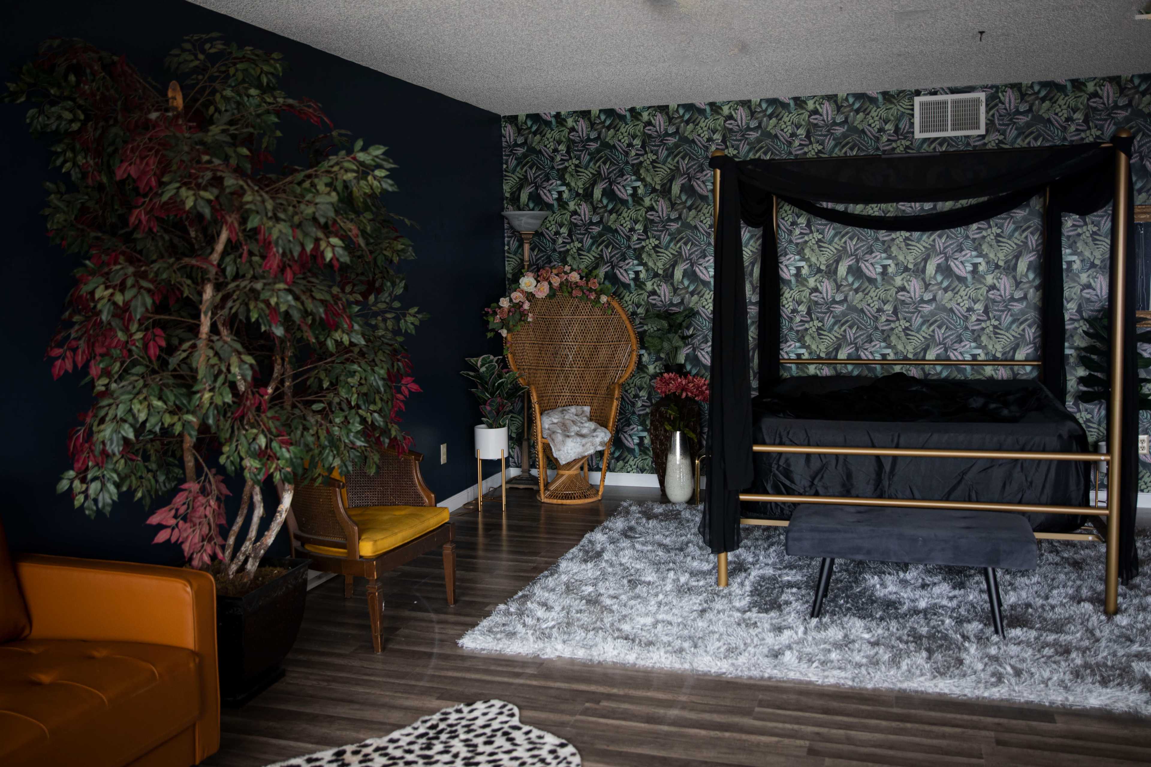 The room features a four-poster bed with a black canopy, surrounded by tropical wallpaper, a large potted plant, and a cozy seating area.