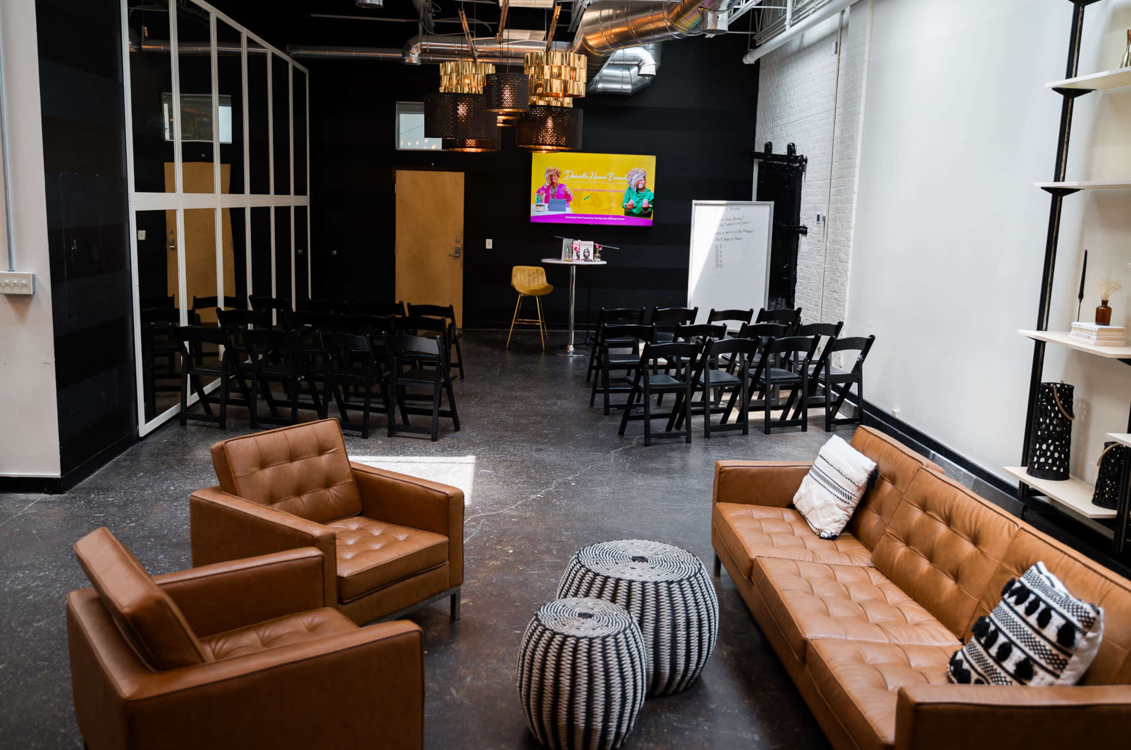 A modern event space with a seating area featuring brown leather sofas and two round tables, facing a stage with a large screen and rows of black folding chairs arranged for an audience.
