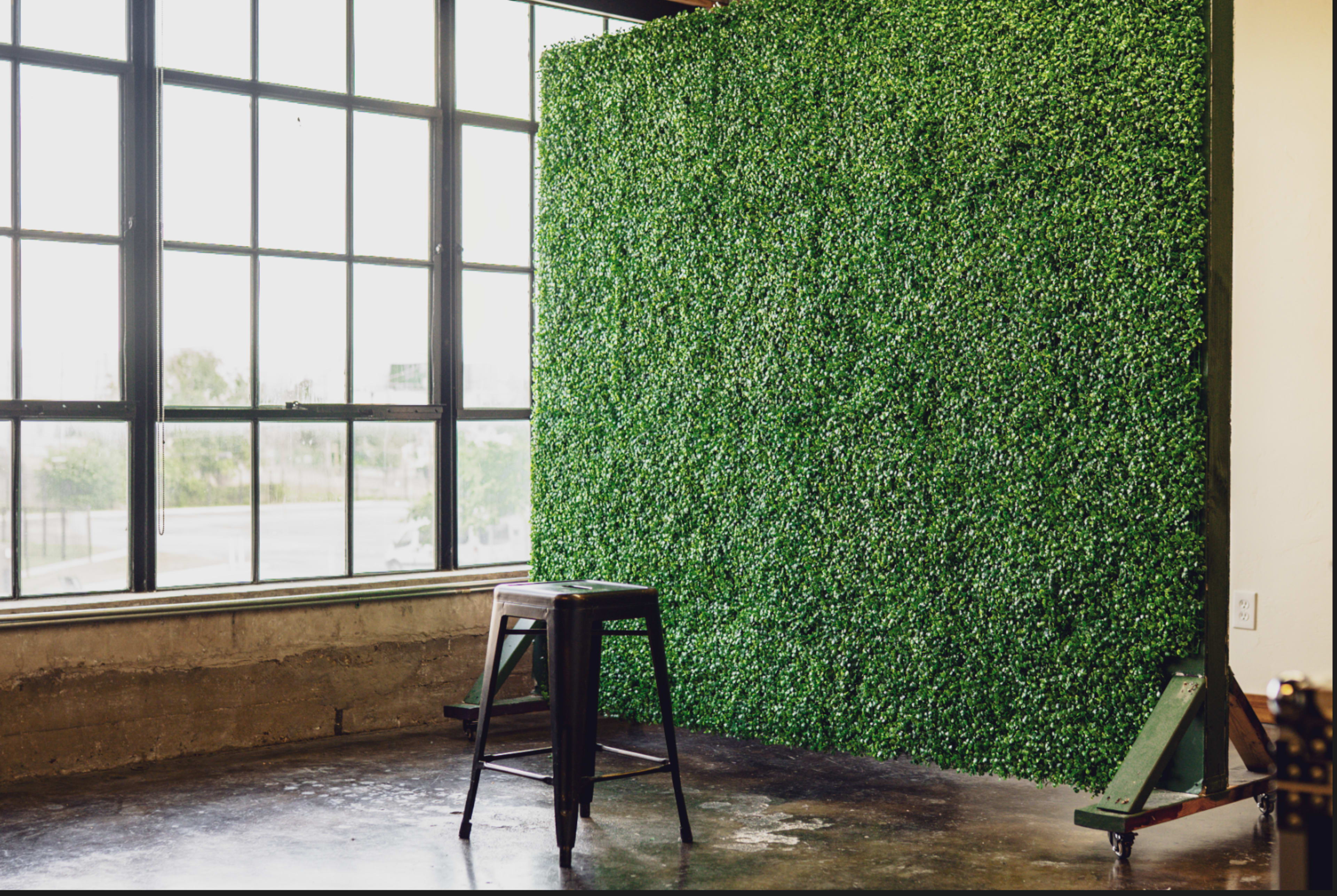 A tall green wall of artificial foliage stands against a windowed backdrop in a room with a polished concrete floor and a metal stool nearby.