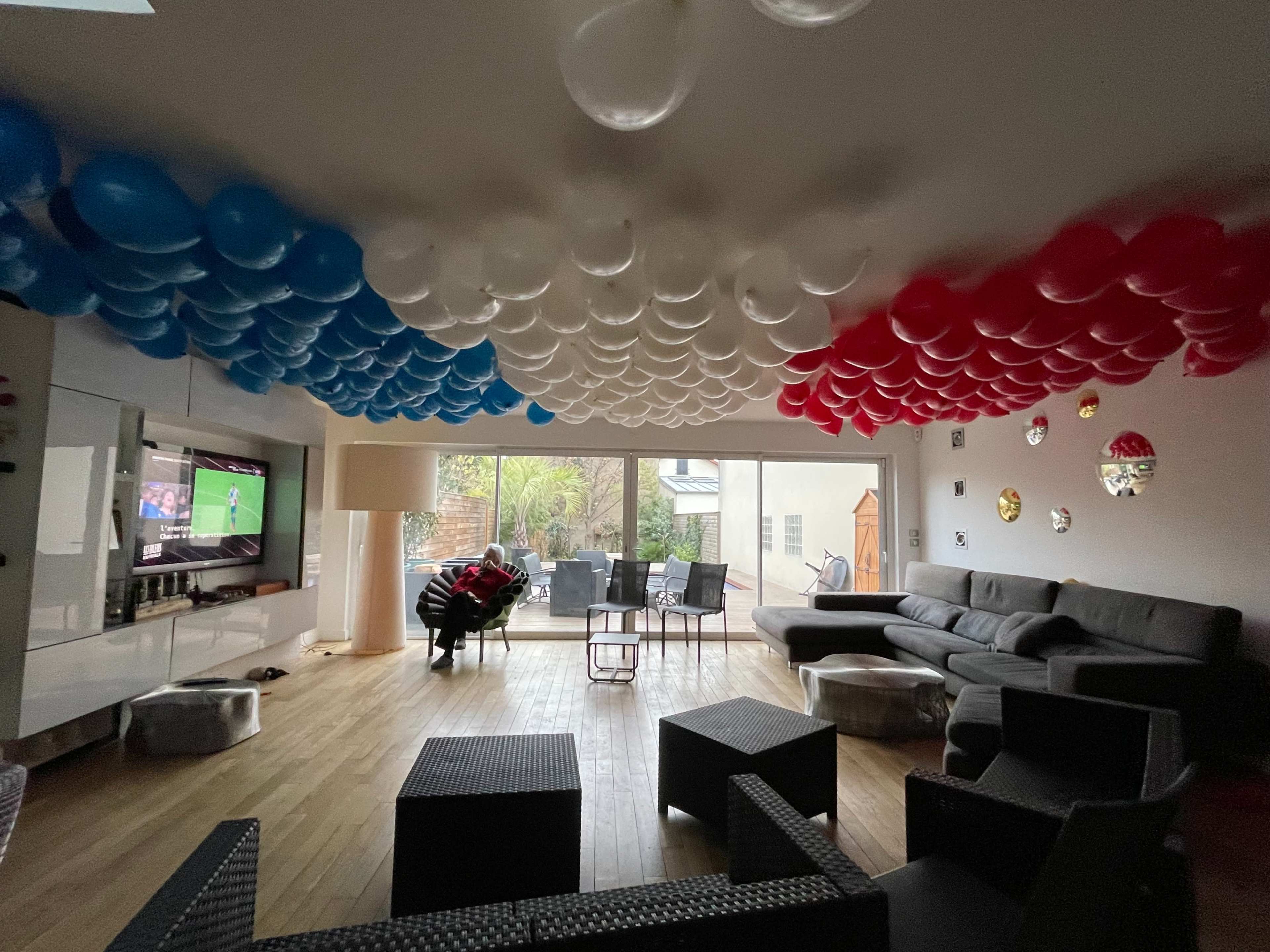 A living room is decorated with red, white, and blue balloons overhead, featuring a television displaying a soccer game and a sectional sofa arranged around a coffee table.