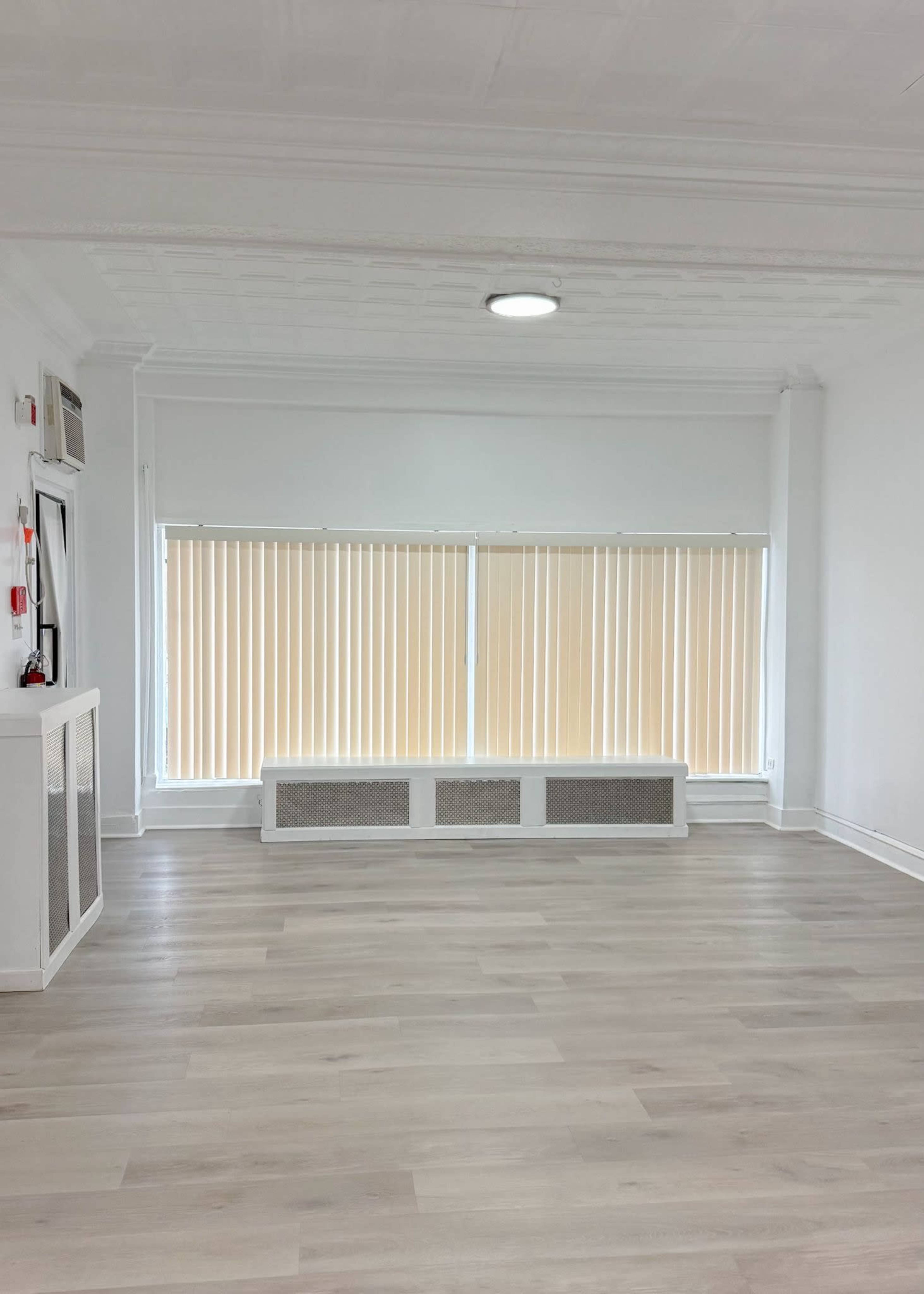 The image shows a spacious, empty room with light-colored wooden flooring and large windows covered by beige vertical blinds.